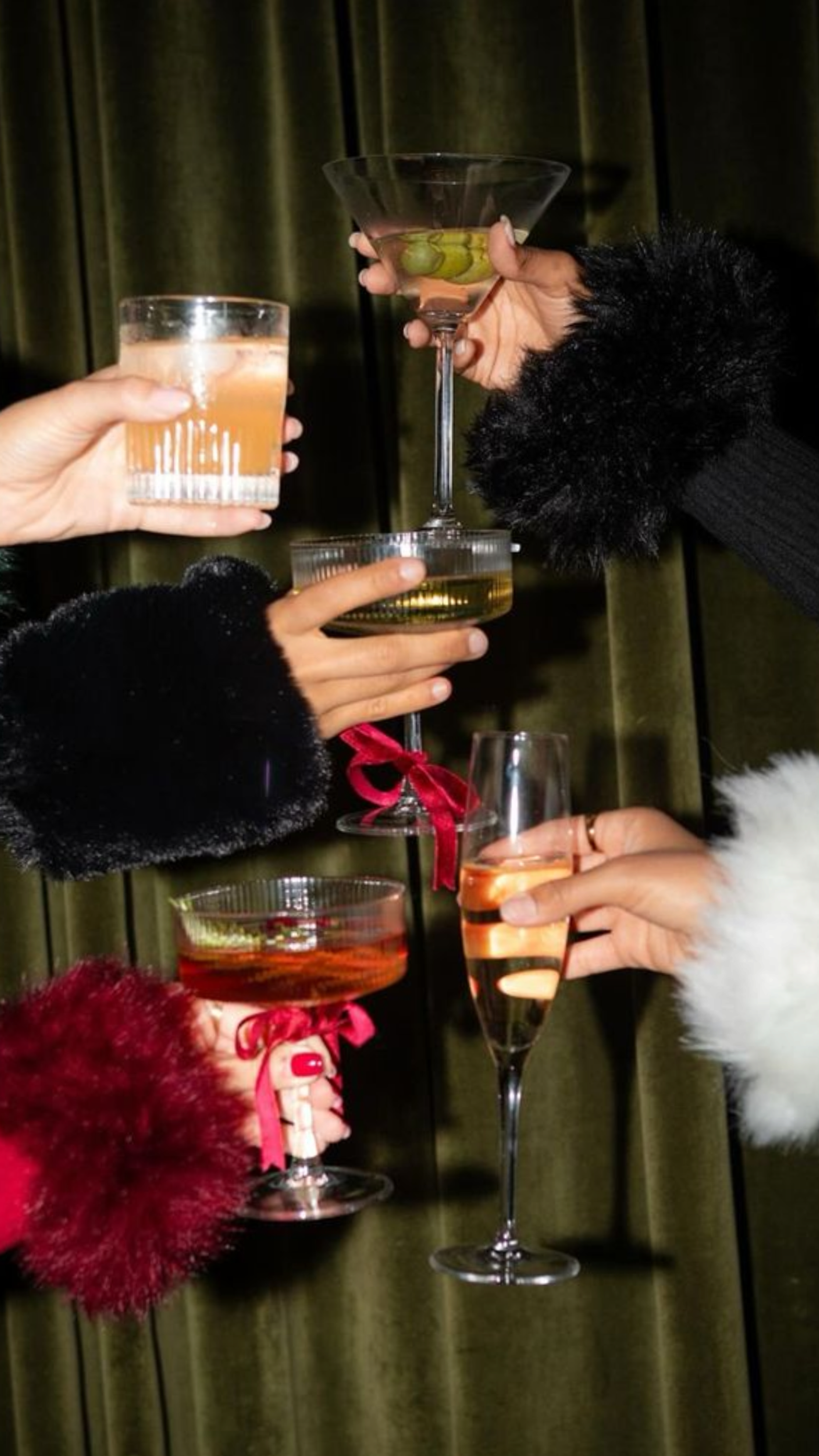 Hands in fluffy faux fur cuffs raising cocktails and coupes against a green velvet curtain