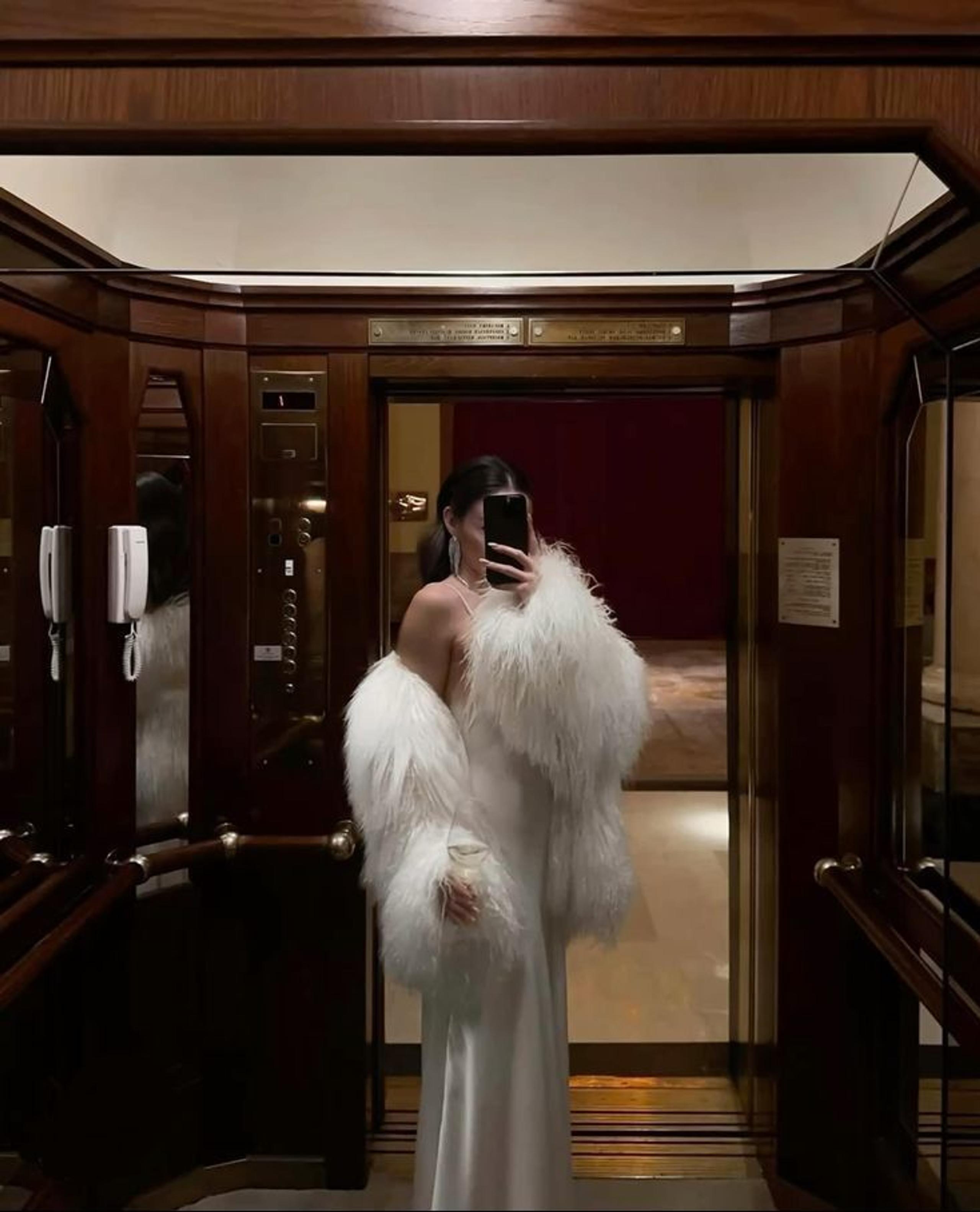 Woman in a white satin slip dress and oversized faux fur stole taking a mirror selfie in a wood-panelled lift