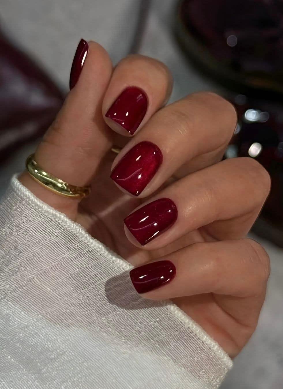 Short square nails painted in a deep glossy red with a gold ring and cream knit sleeve
