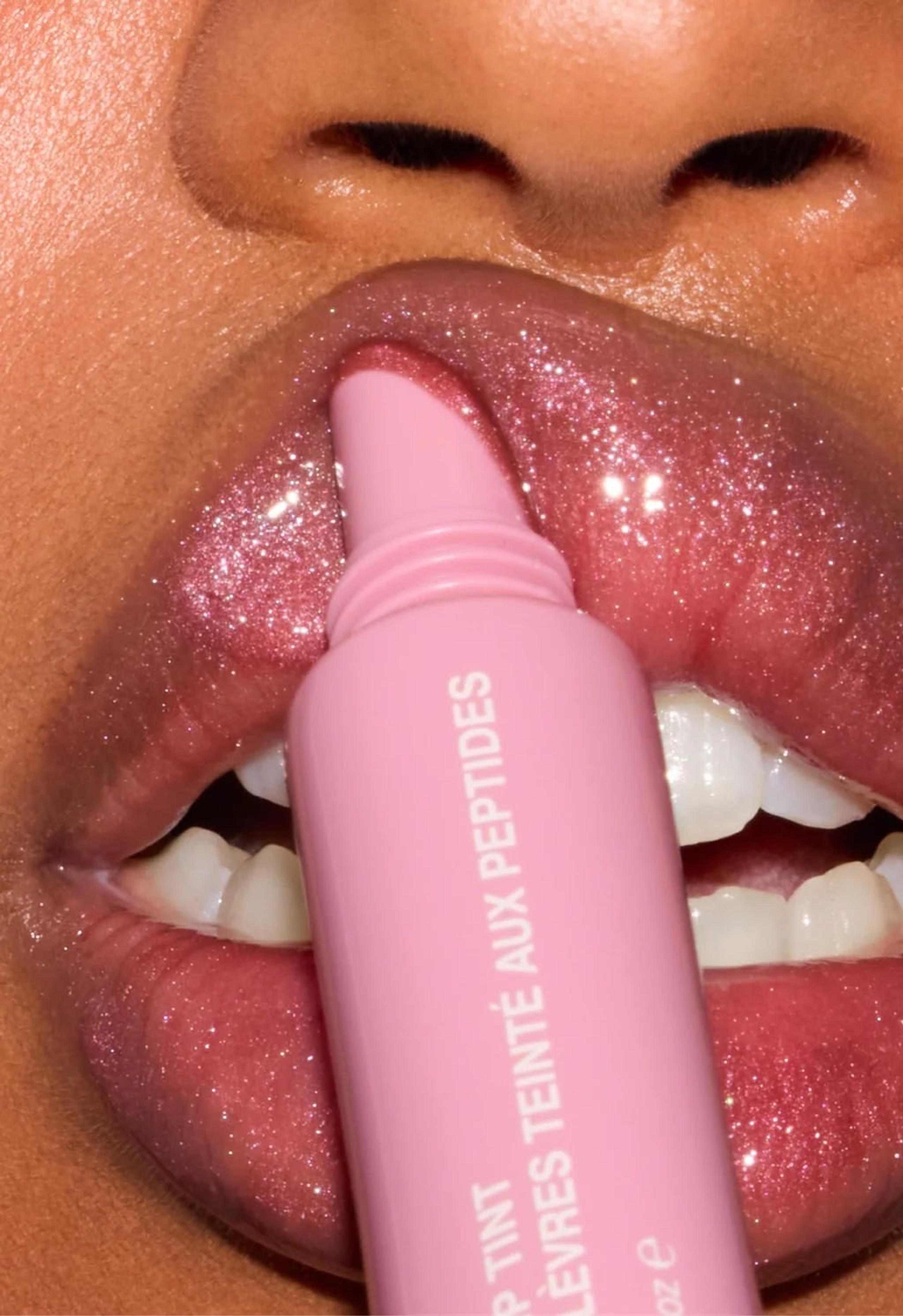 Close-up of lips wearing a glossy berry lip colour, with lipstick bullets and gloss wands arranged beside them