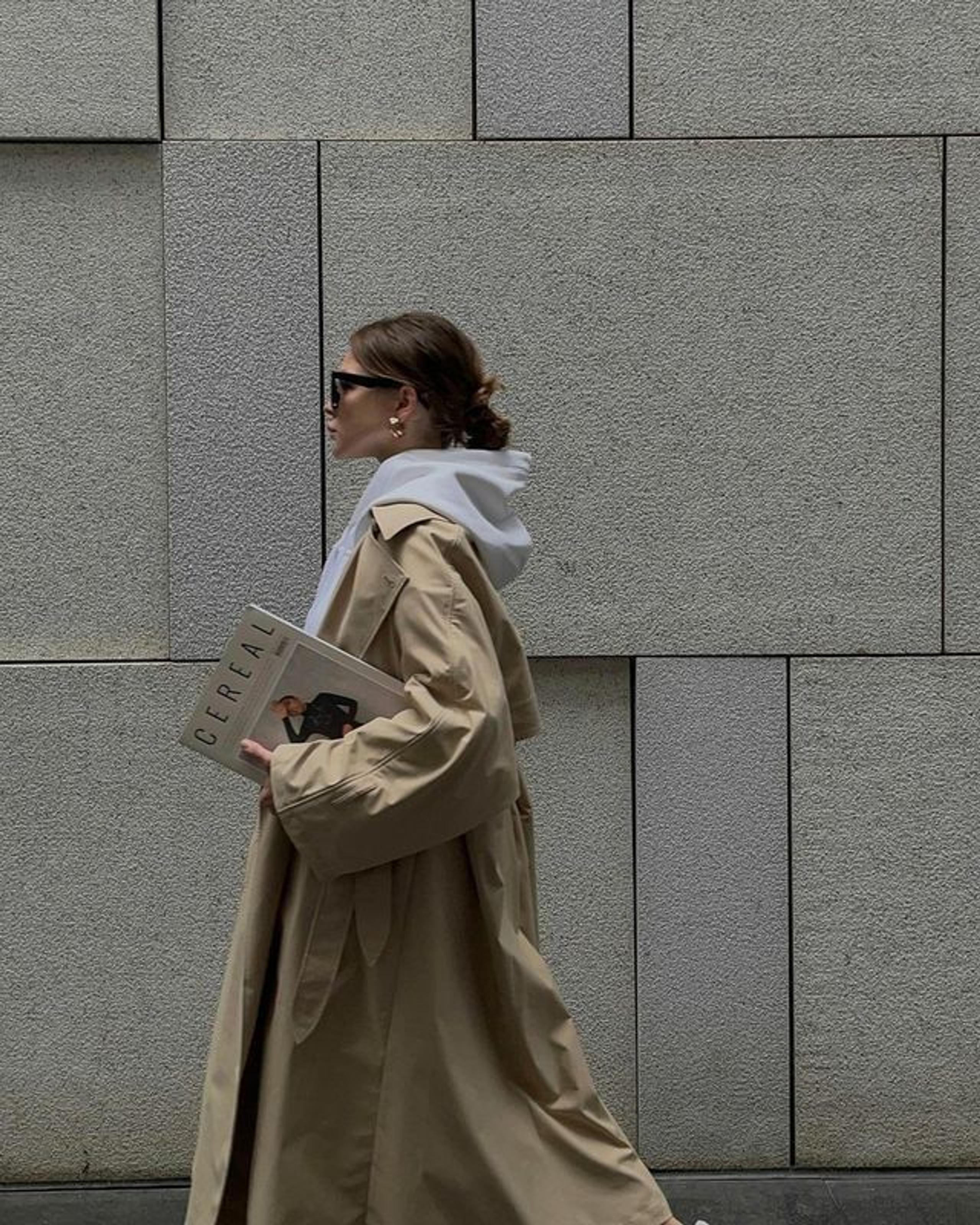 Woman in a long beige trench over a grey hoodie carrying a magazine past a concrete wall