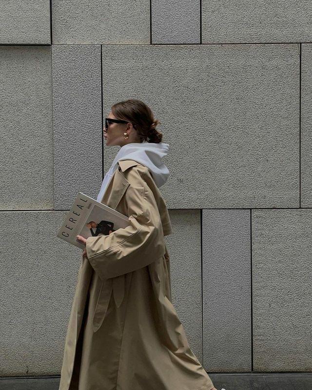 Woman in a long beige trench over a grey hoodie carrying a magazine past a concrete wall