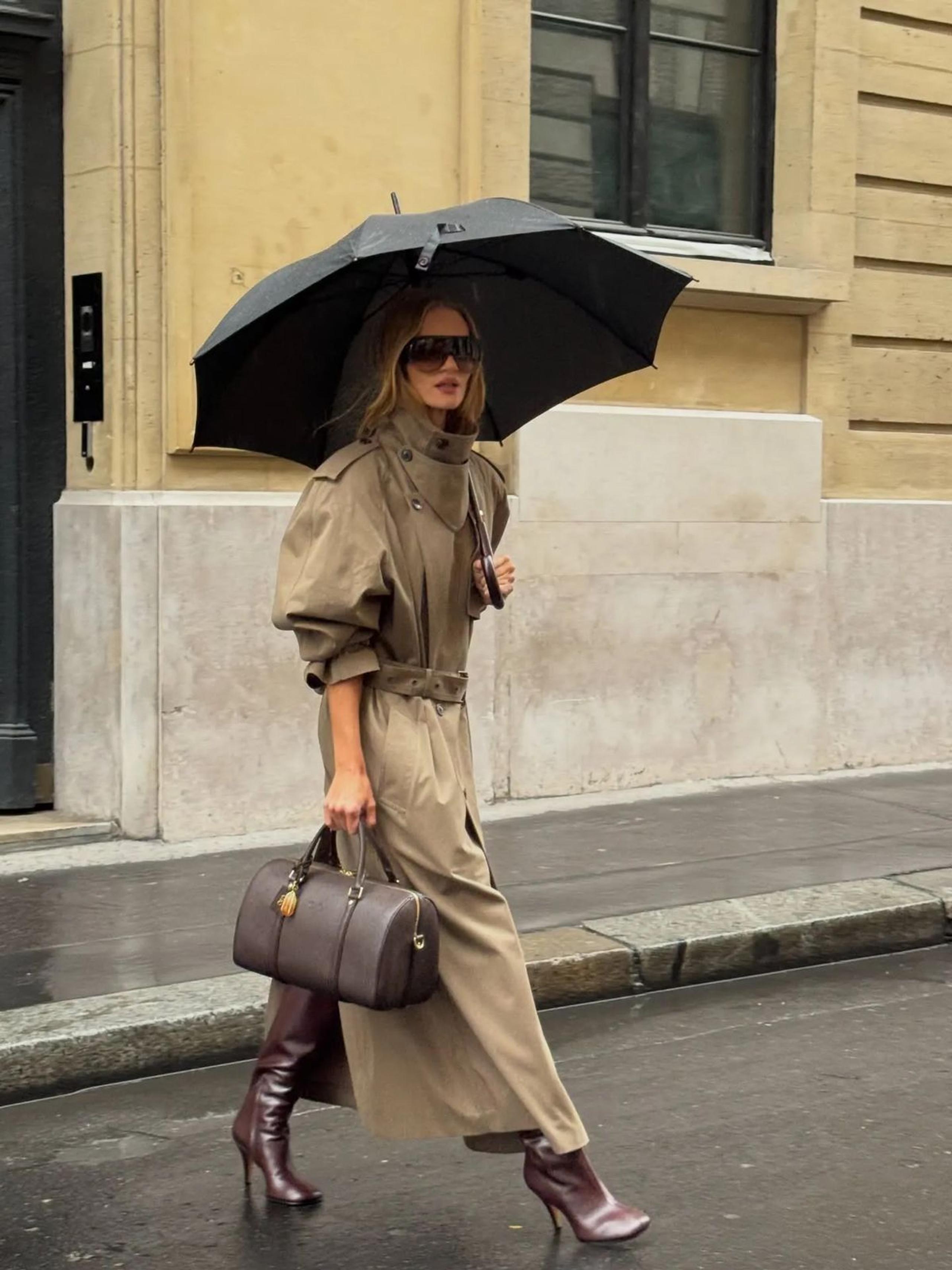 Woman in a belted khaki trench and burgundy heeled boots walking under a black umbrella on a rainy street