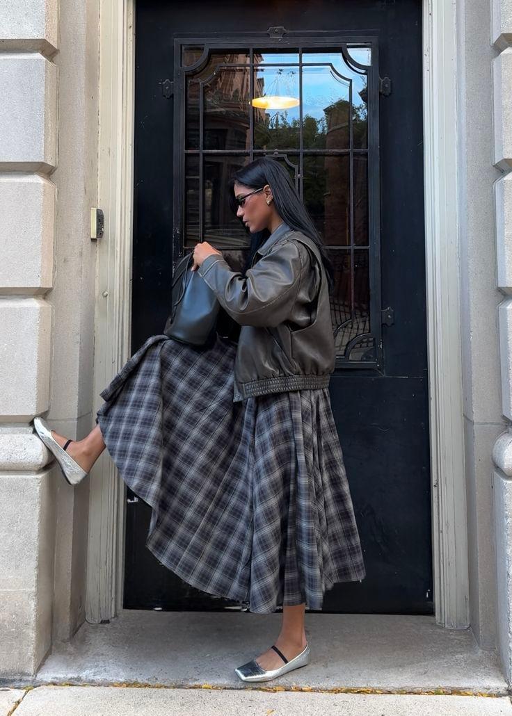 Woman in a full grey plaid midi skirt, brown leather bomber and silver ballet flats in a doorway