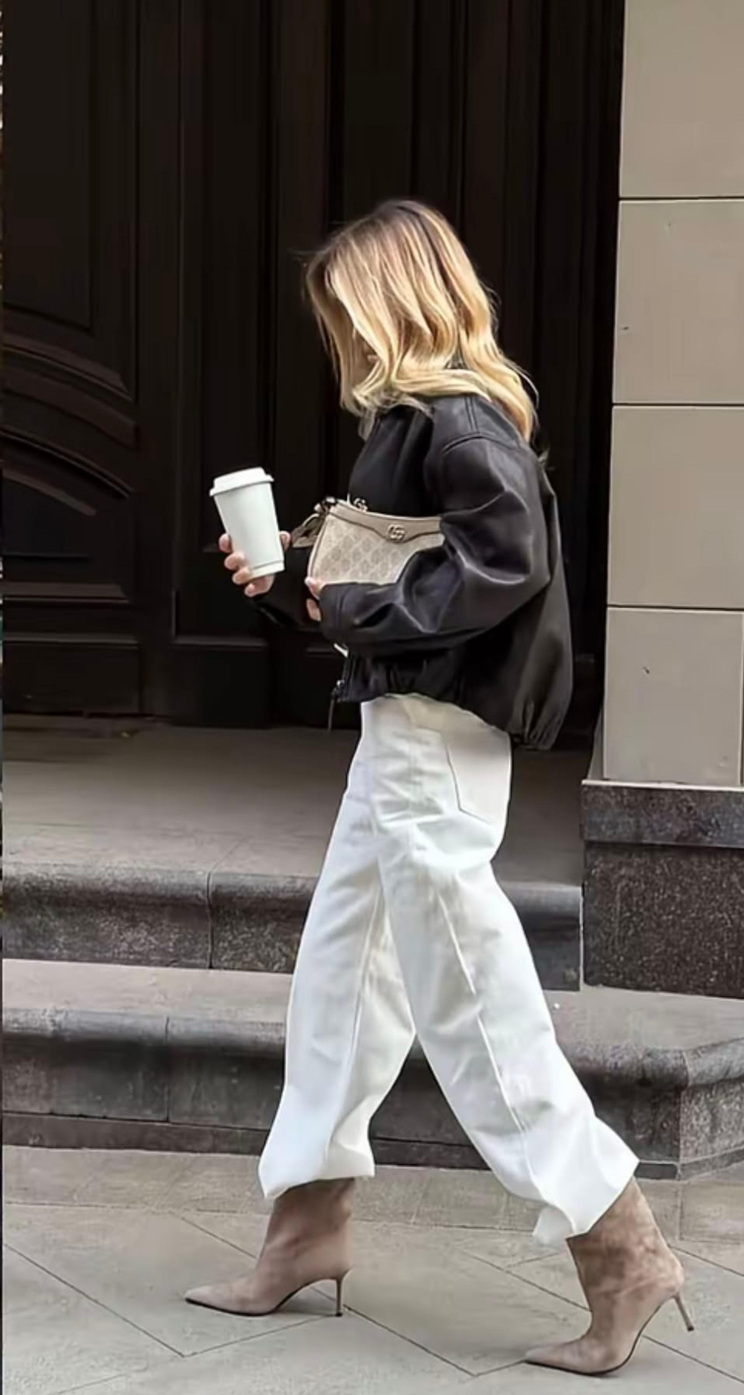 Woman in white wide-leg jeans and a cropped black leather jacket carrying a coffee