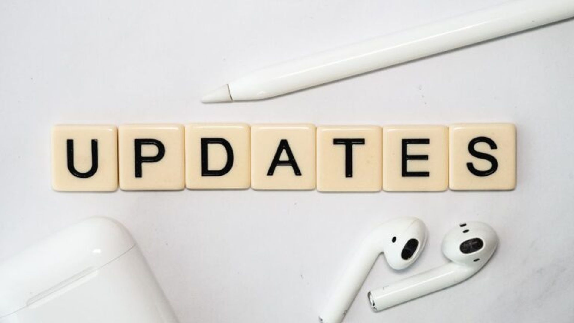 The word "UPDATES" spelled out in letter tiles, with a white stylus and wireless earbuds with their charging case on a white surface.