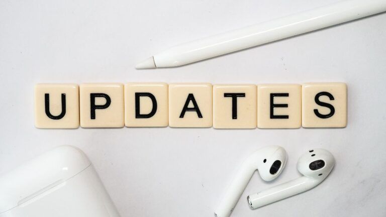 The word "UPDATES" spelled out in letter tiles, with a white stylus and wireless earbuds with their charging case on a white surface.