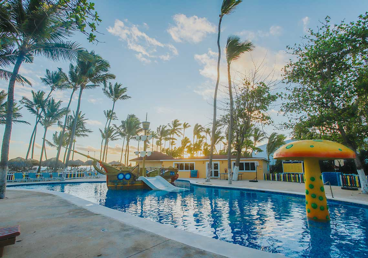 Kids pool with a pirate ship water slide and mushroom fountain at the Grand Bavaro Princess resort in Punta Cana, surrounded by palm trees.
