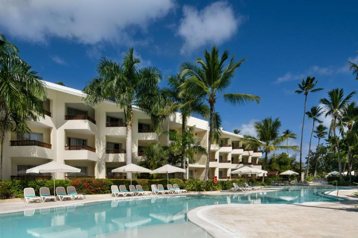 View of the swimming pool area at the Impressive Punta Cana resort, featuring white hotel buildings, palm trees, sun loungers, and parasols under a blue sky.