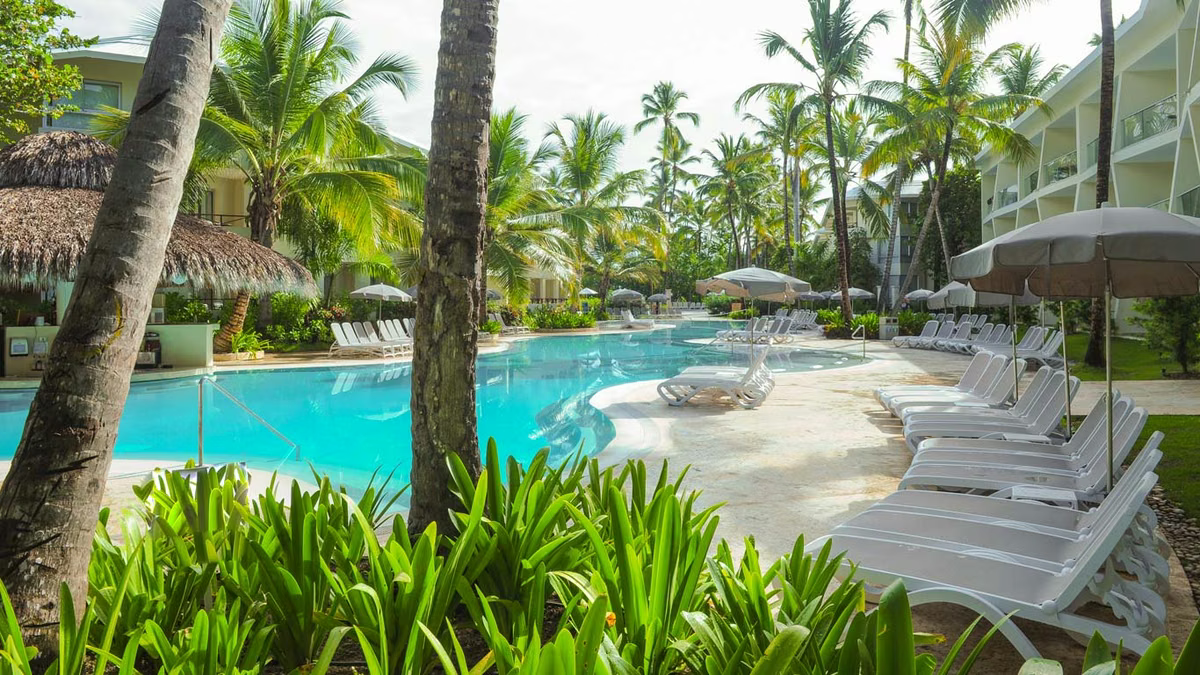 Resort swimming pool area surrounded by palm trees, sun loungers with umbrellas, and modern hotel buildings at Impressive Punta Cana in the Dominican Republic.
