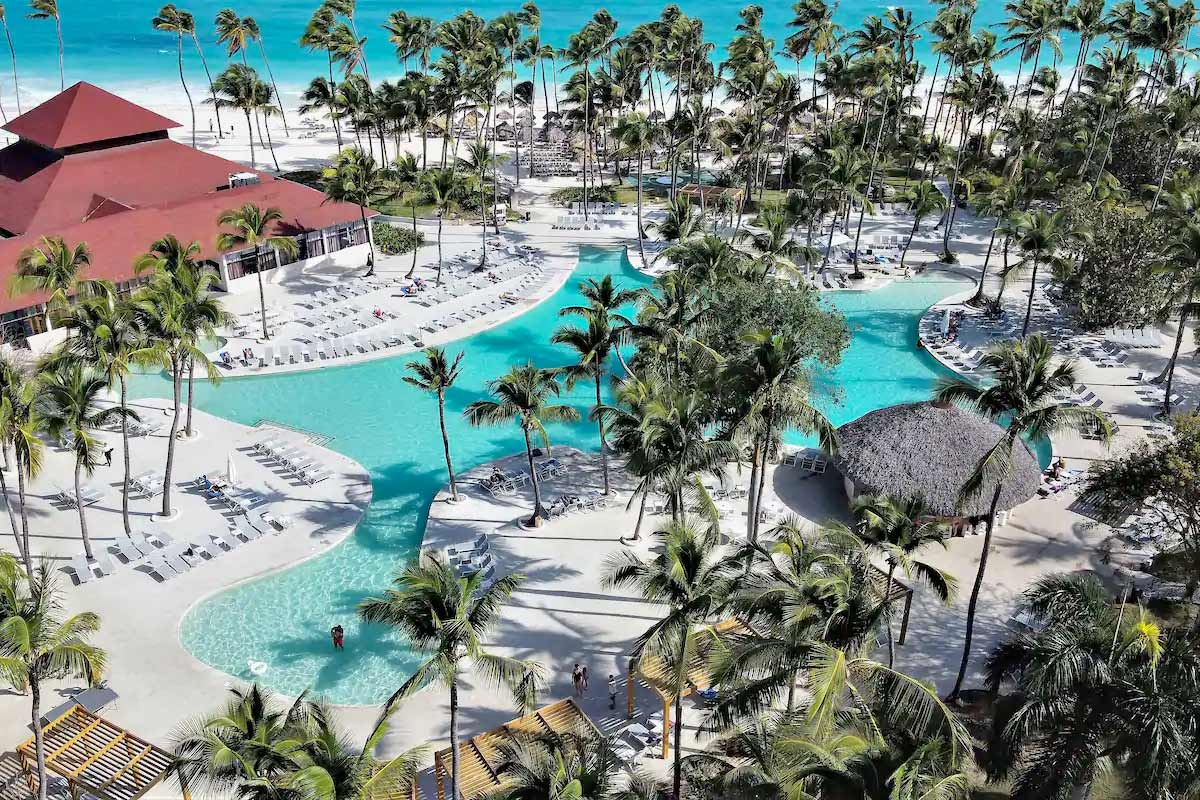 Aerial view of the large outdoor swimming pool and palm trees at the Grand Bavaro Princess resort in Punta Cana, showing sun loungers, resort buildings, and the nearby beach.