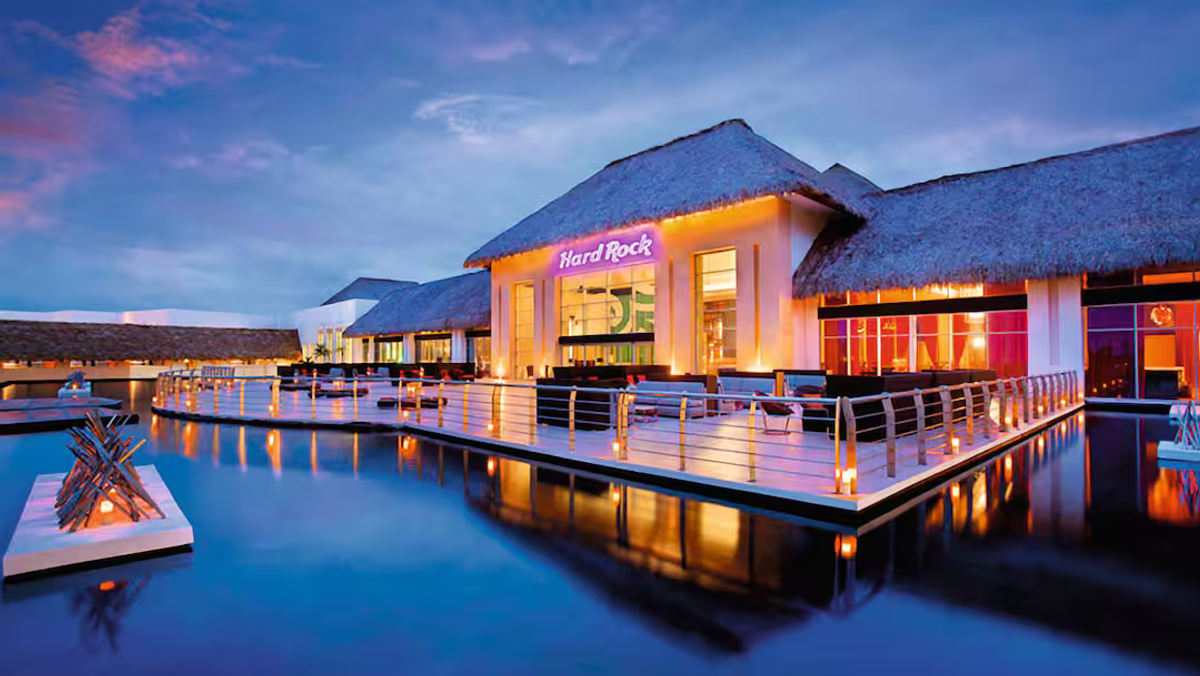 Illuminated exterior view of the Hard Rock Hotel and Casino Punta Cana featuring modern architecture, thatched roofs, an outdoor terrace lounge, and water reflections at dusk.