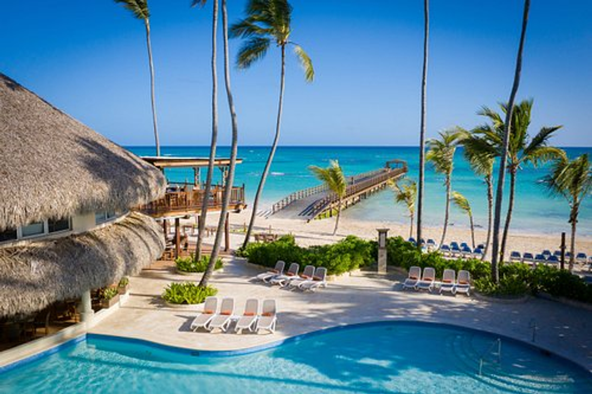 Tropical resort view from a terrace overlooking a swimming pool, palm trees, a thatched-roof building, the beach, and a long wooden pier leading into the turquoise Caribbean Sea at Impressive Punta Cana.