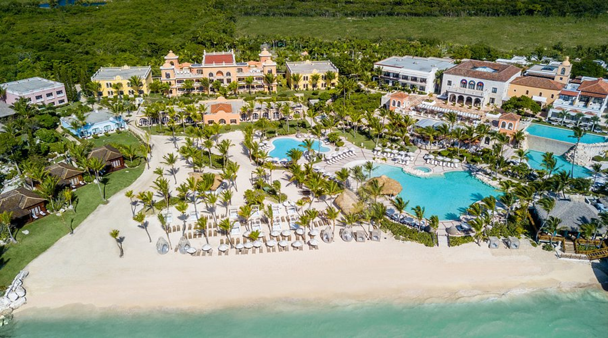 Aerial view of Sanctuary Cap Cana resort, showcasing its architecture, sprawling pools, palm trees, sun loungers on the sandy beach, and clear turquoise water[cite: 7].