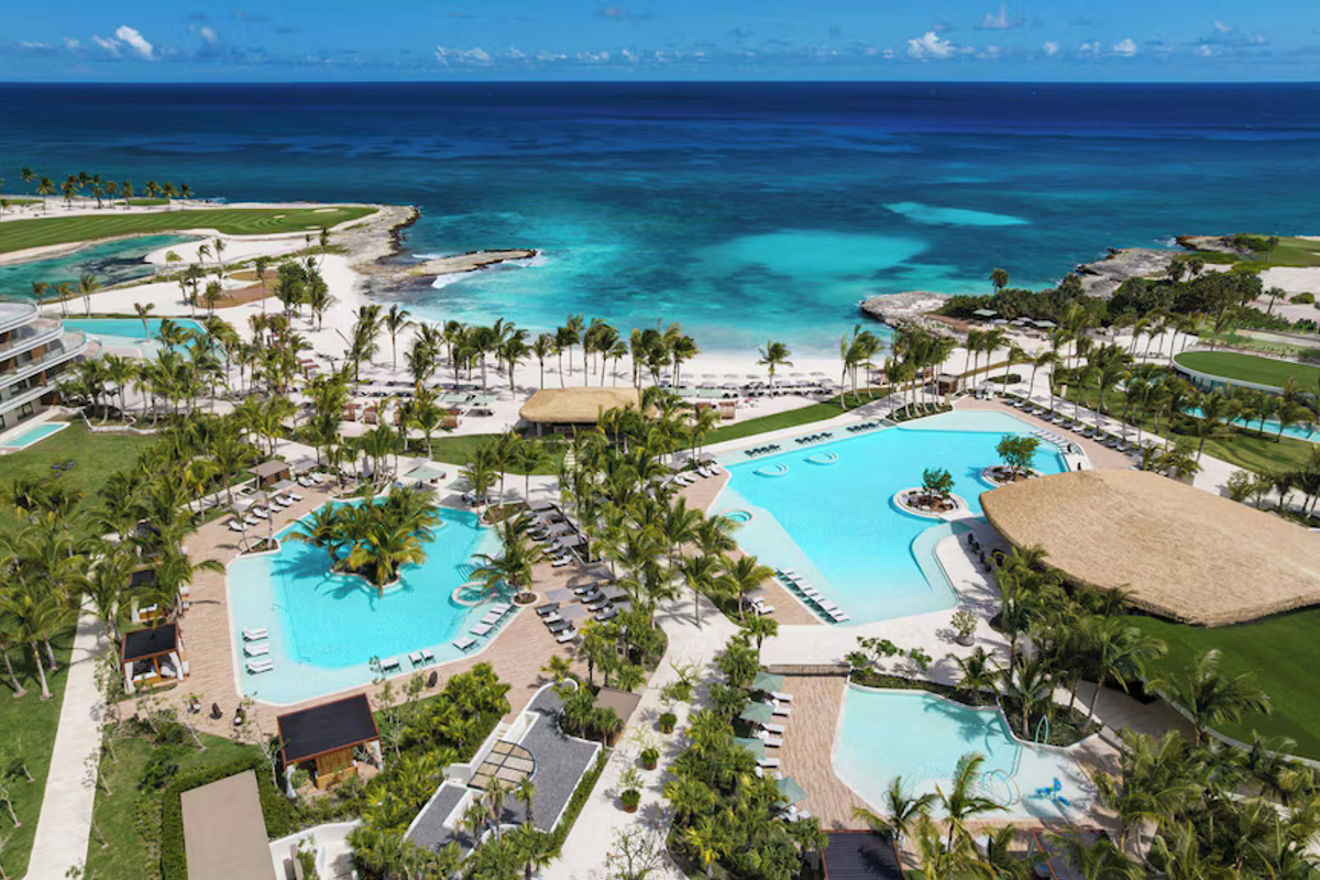 High-angle aerial view of the swimming pool areas, lush palm trees, and pristine white-sand beach overlooking the turquoise ocean at The St. Regis resort in Punta Cana.