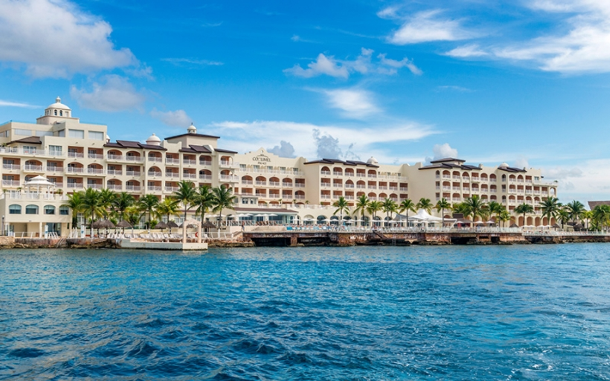 Exterior view of the Barceló Bávaro Palace resort facade seen from the sea, featuring multi-story white buildings, palm trees, and coastal waters.