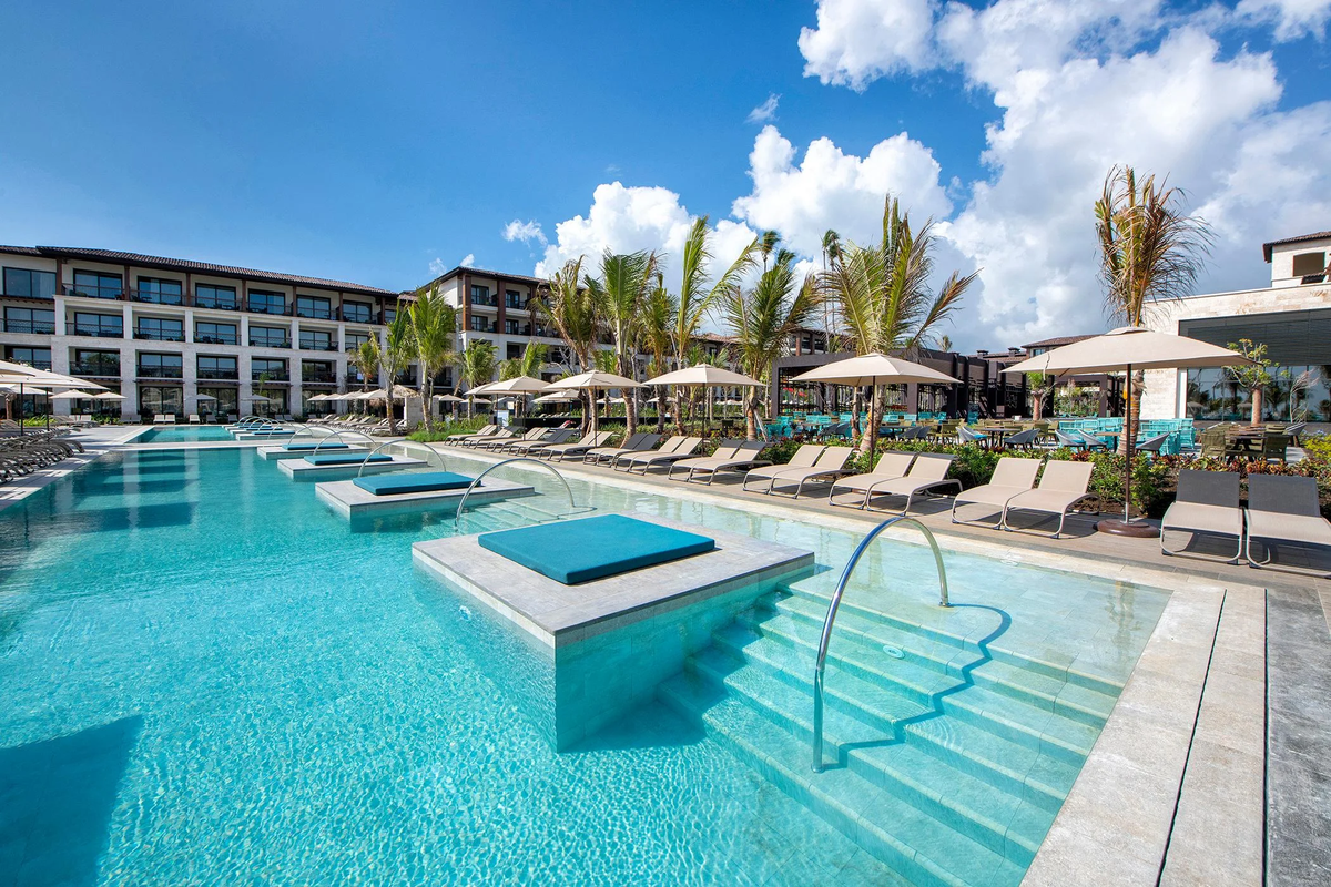 Ground-level exterior view of a large swimming pool at the Lopesan Costa Bávaro Resort, Spa & Casino, featuring submerged sun pads, lounge chairs, umbrellas, palm trees, and resort buildings under a partly cloudy blue sky.
