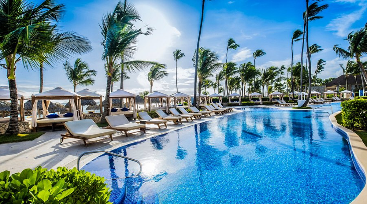 Ground-level view of a swimming pool at the Majestic Colonial Punta Cana, surrounded by lounge chairs, cabanas, and tall palm trees under a bright sunny sky[cite: 9].