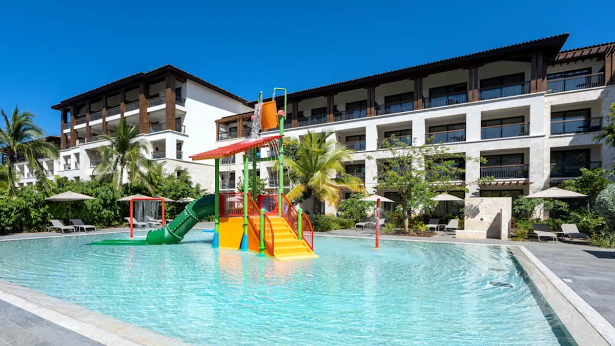 Exterior view of a children's splash pool at the Lopesan Costa Bávaro Resort, Spa & Casino, featuring a colorful water slide, a dumping bucket structure, palm trees, and resort buildings under a clear blue sky[cite: 10].