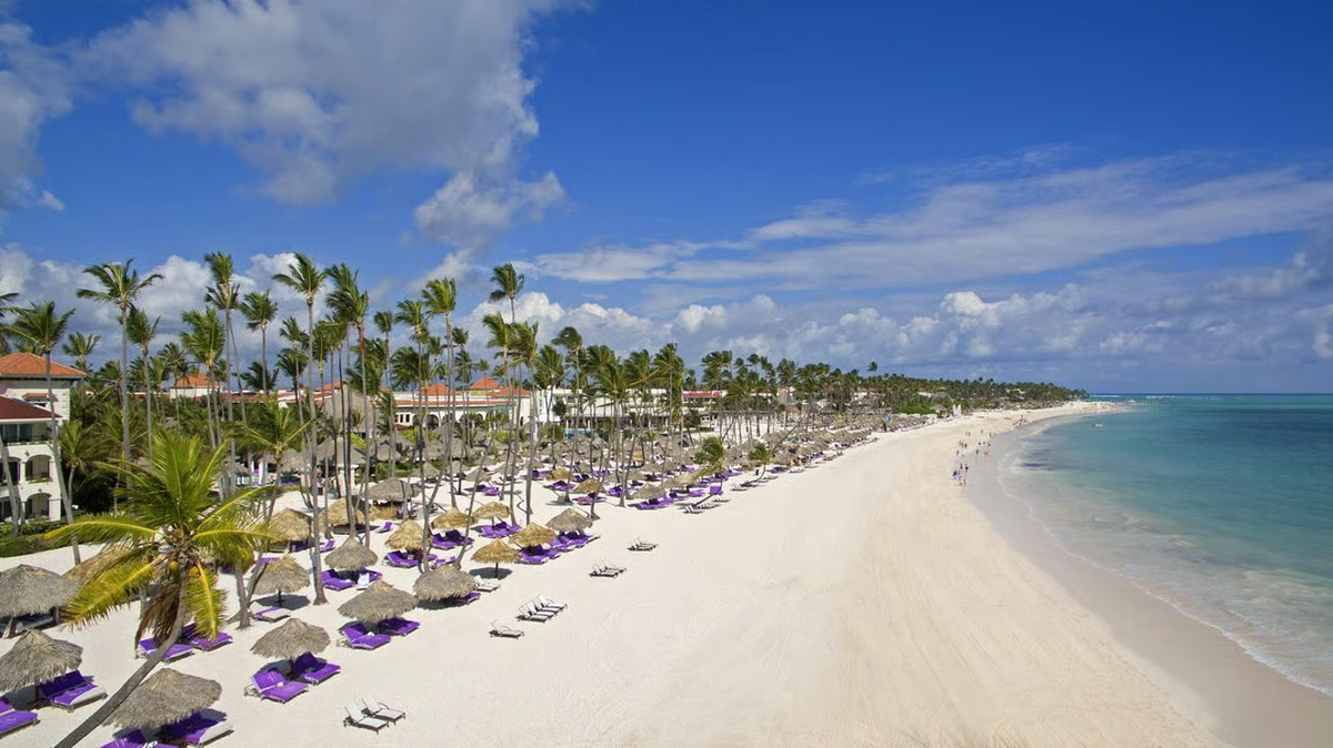 Scenic view of the pristine white sand beach lined with palm trees, palapas, and lounge chairs along the turquoise ocean at Paradisus Palma Real Golf & Spa Resort in Punta Cana.