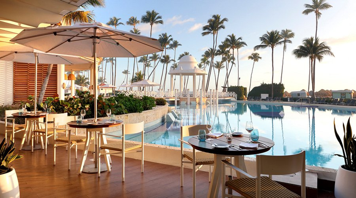 Outdoor dining terrace at Paradisus Palma Real Golf & Spa Resort in Punta Cana, overlooking the swimming pool and palm trees, with tables, chairs, and umbrellas under a blue sky.