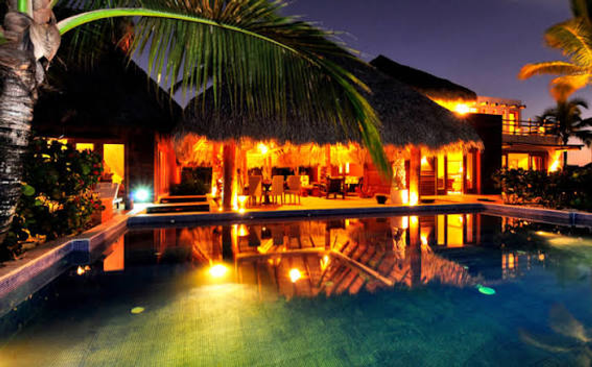 Illuminated exterior view of a luxury villa at Villas Caleton 51 featuring a private swimming pool, warm lighting reflecting on the water, and traditional thatched-roof architecture at night.