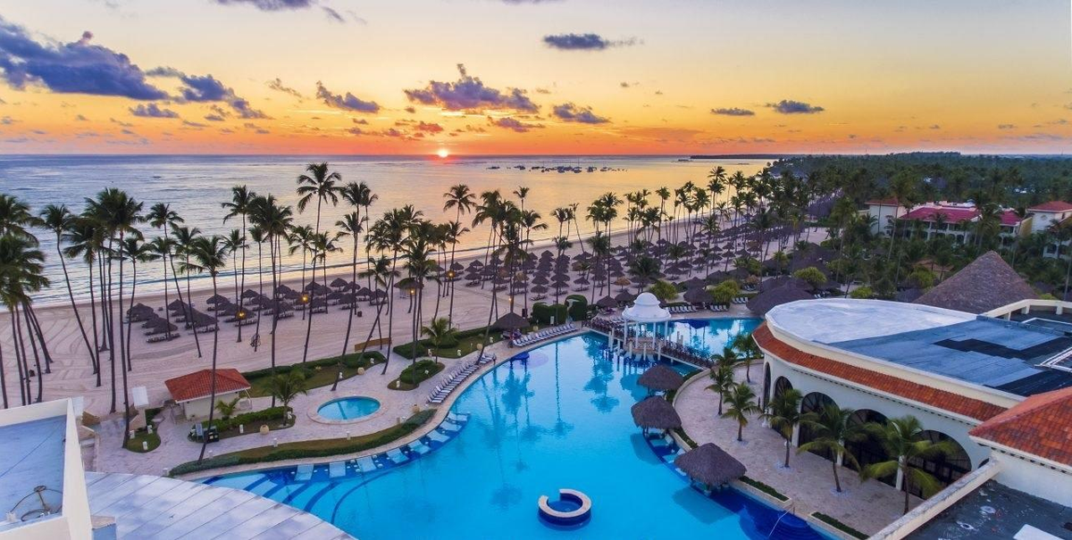 Aerial view of the Paradisus Palma Real Golf & Spa Resort in Punta Cana at sunset, showing the swimming pools, palm trees, beach with palapas, and the ocean.