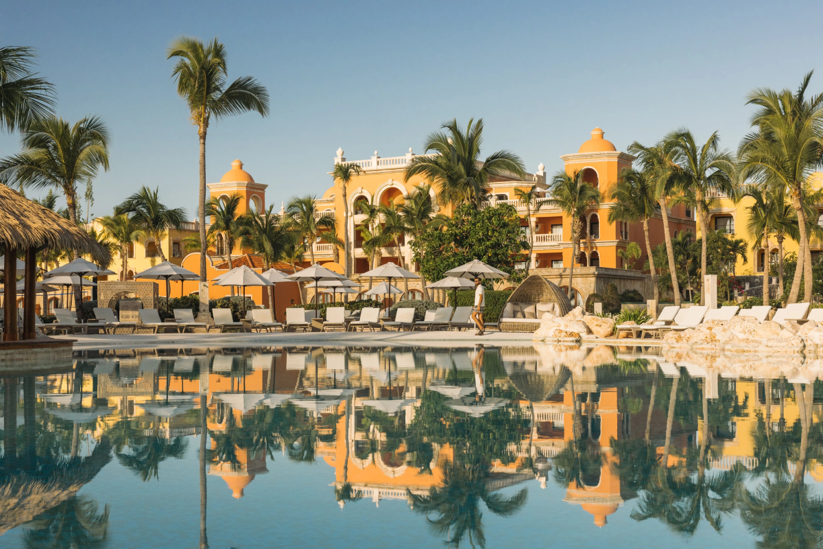 Exterior view of the Spanish colonial-style architecture at Sanctuary Cap Cana, featuring warm orange buildings, palm trees, and loungers surrounding a large swimming pool that reflects the resort scenery.