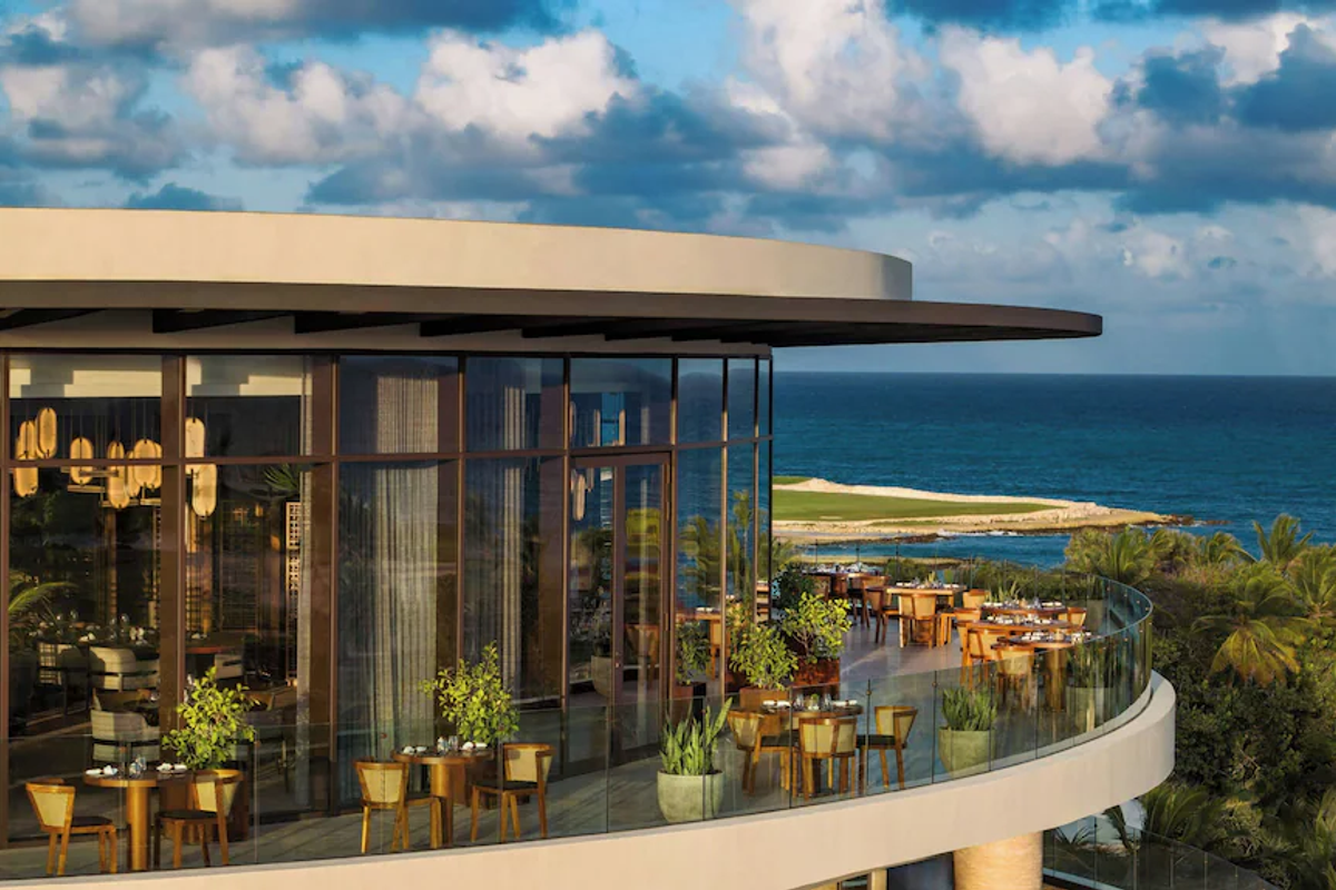 Exterior view of a curved terrace and restaurant dining area with glass railings at The St. Regis resort in Punta Cana, overlooking the ocean under a cloudy sky.