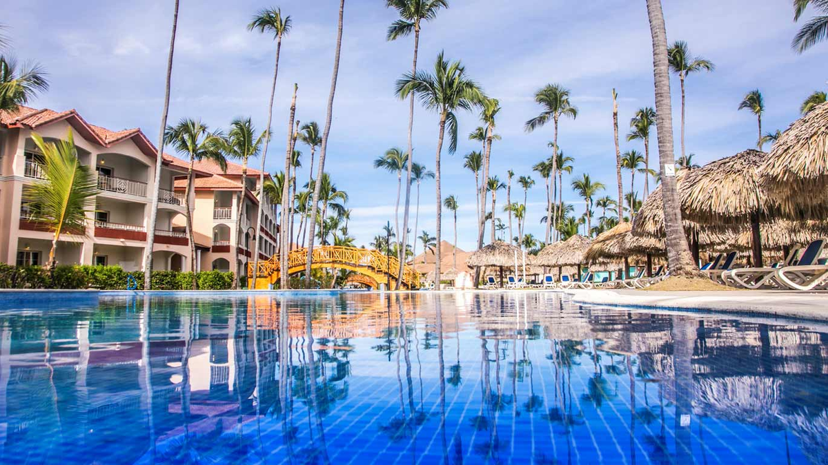 Ground-level view of a clear swimming pool reflecting palm trees, resort buildings, and a bridge at the Majestic Colonial Punta Cana, surrounded by tropical vegetation and thatched umbrellas.