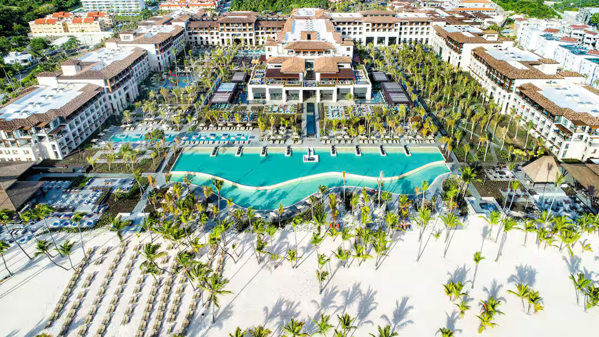 Aerial view of the Lopesan Costa Bávaro Resort, Spa & Casino, showing its large turquoise swimming pools, palm trees, expansive white-sand beach with sun loungers, and resort buildings under daylight.
