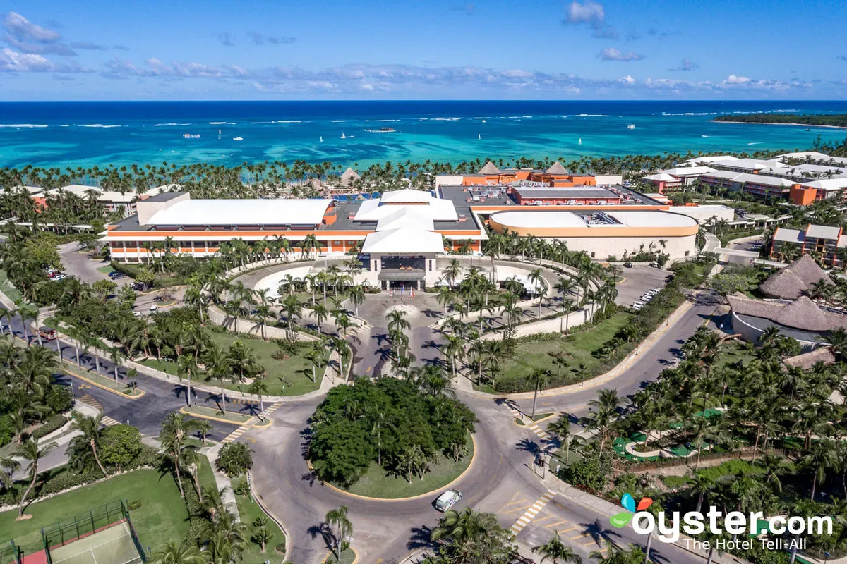 Aerial view of the entrance and main buildings of the Barceló Bávaro Palace resort, surrounded by lush landscaping and roundabouts, with the ocean in the background.