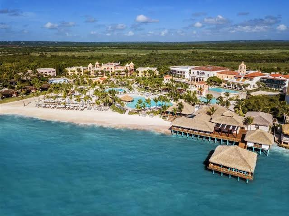 Aerial view of Sanctuary Cap Cana resort, showing its Spanish colonial-style buildings, swimming pools, palm trees, beach, and overwater structures along the turquoise sea
