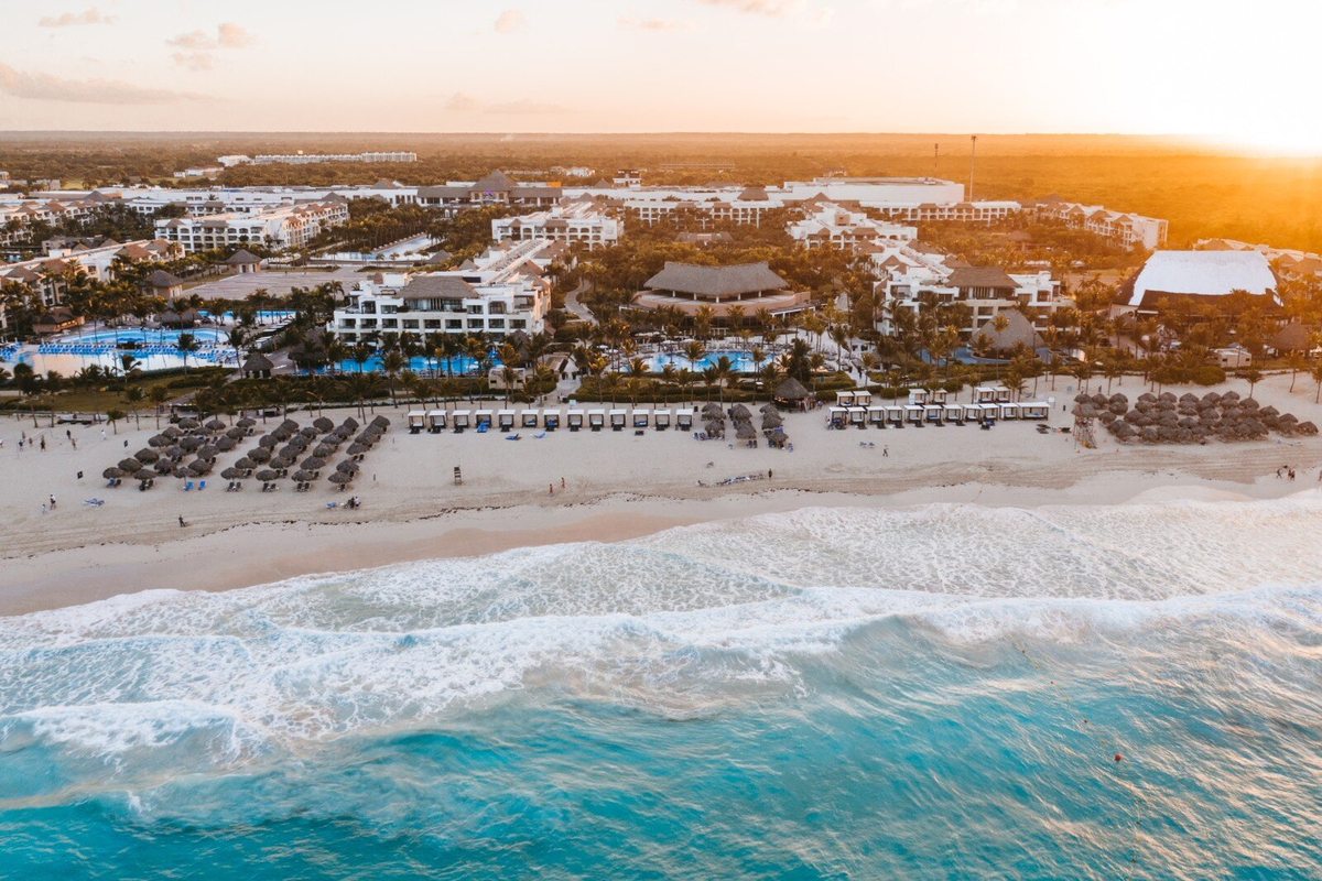 Aerial drone view of the Hard Rock Hotel and Casino Punta Cana during sunset, showcasing the sprawling resort property, lush palm trees, pristine white sand beach, and turquoise ocean waves.