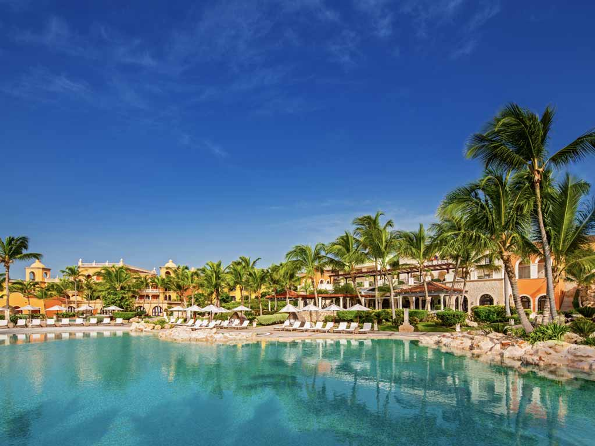 Exterior view of a large swimming pool at Sanctuary Cap Cana, surrounded by lounge chairs, palm trees, and the resort's buildings under a bright blue sky