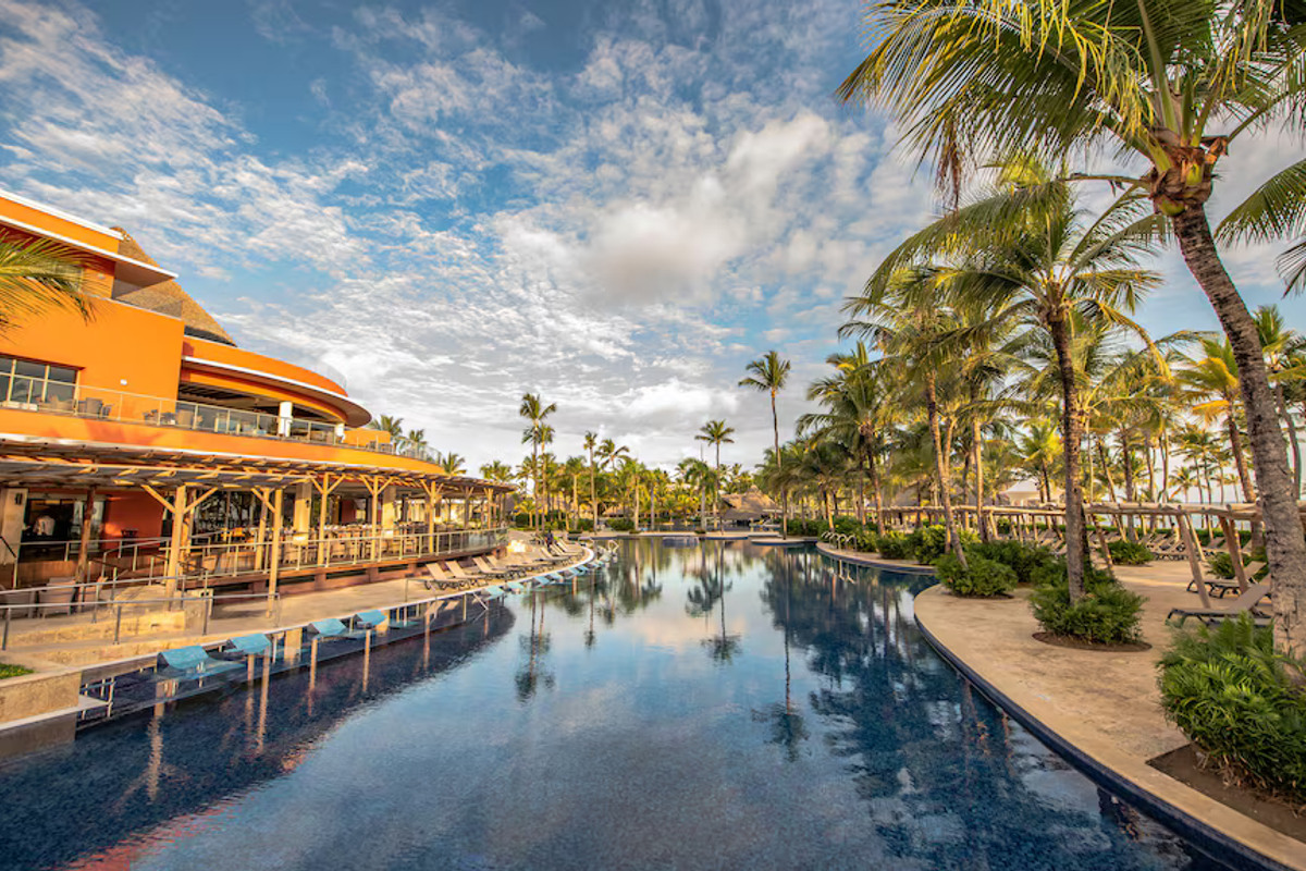 Exterior view of a long swimming pool stretching alongside resort buildings and towering palm trees under a bright cloudy sky.