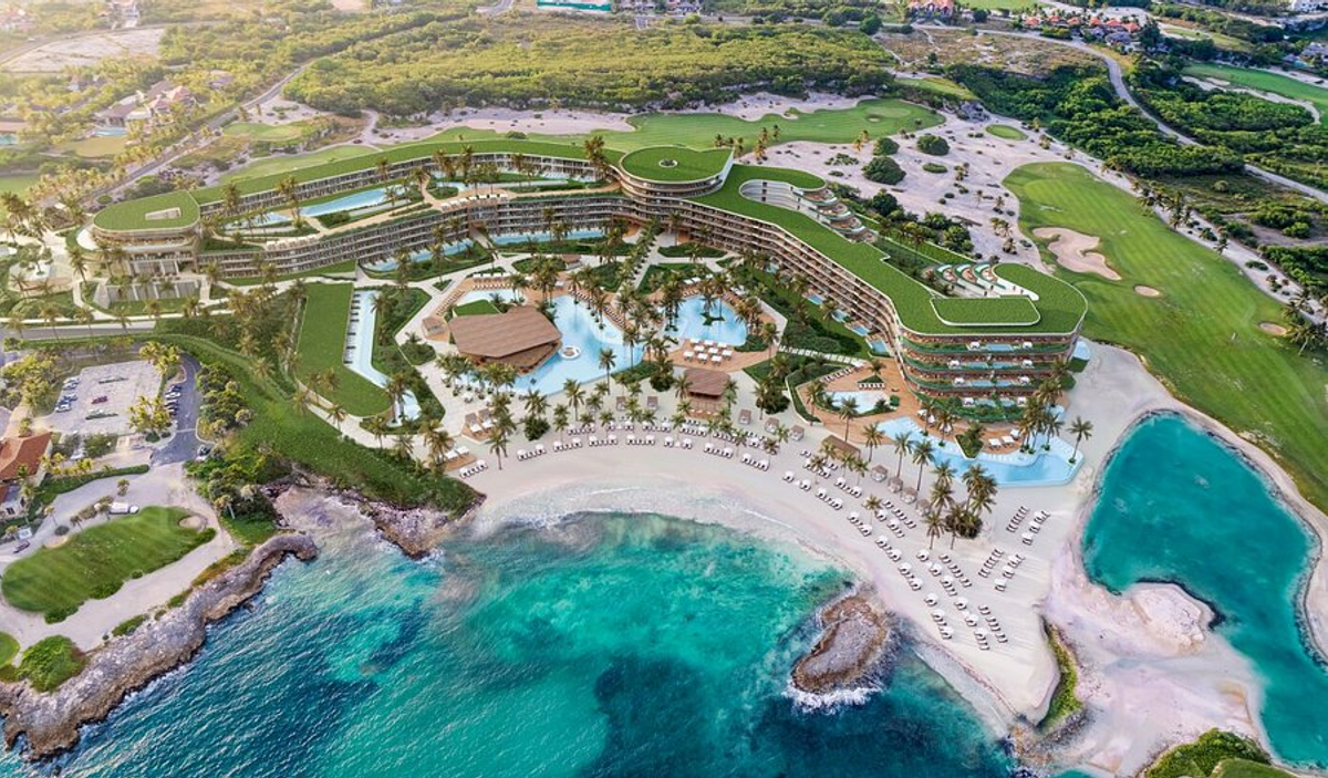 Aerial view of The St. Regis resort in Punta Cana, showing its sprawling multi-wing design with green rooftops, large swimming pool areas, and palm trees along a sandy beach next to turquoise ocean waters.