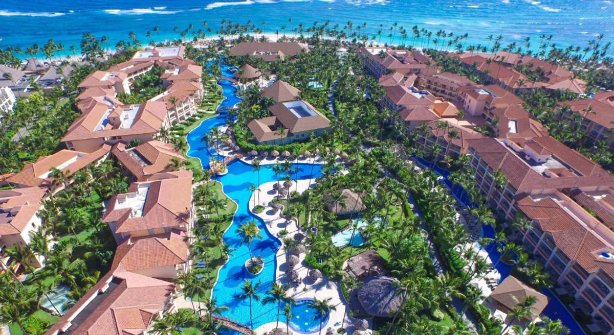 Aerial view of the Majestic Colonial Punta Cana resort, showcasing its long winding pool canals, buildings with red tile roofs surrounded by tropical vegetation, and the beach in the background.