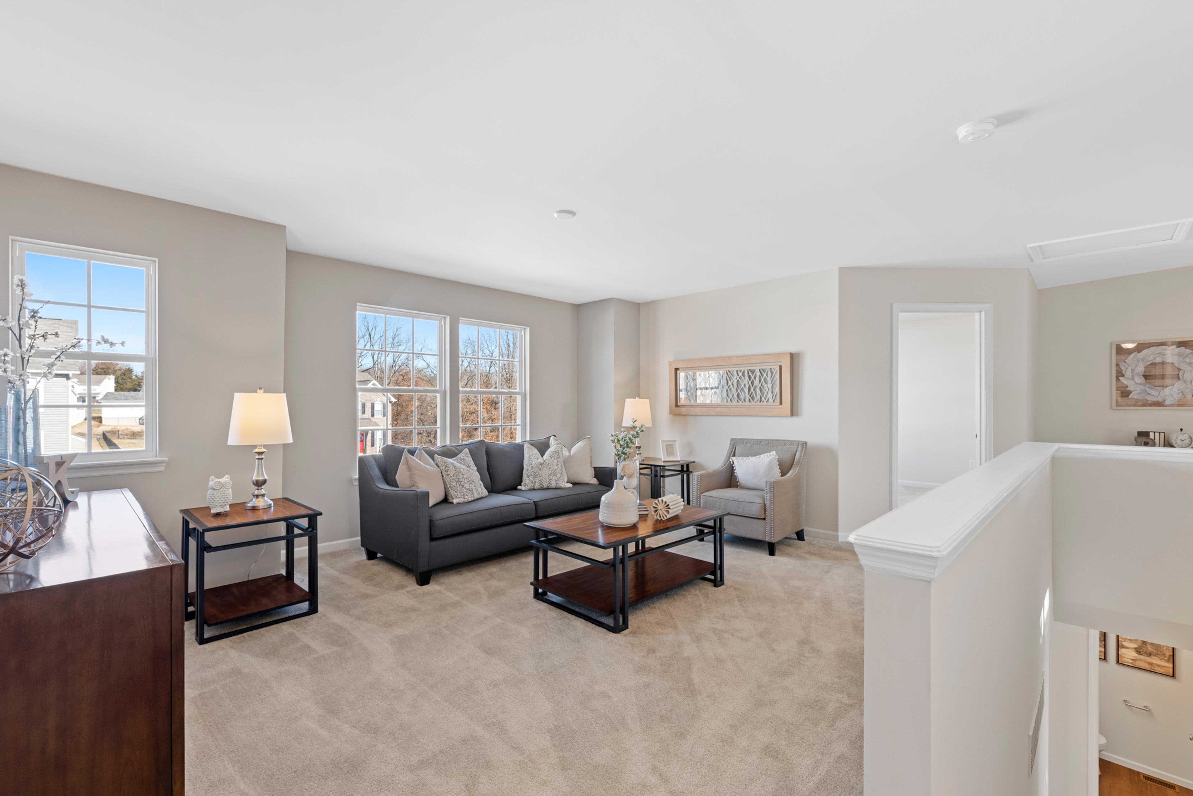 A spacious, light-filled living area featuring neutral tones, plush carpeting, and comfortable seating arranged around a sleek coffee table. Large windows bring in natural light, creating a warm and inviting atmosphere.