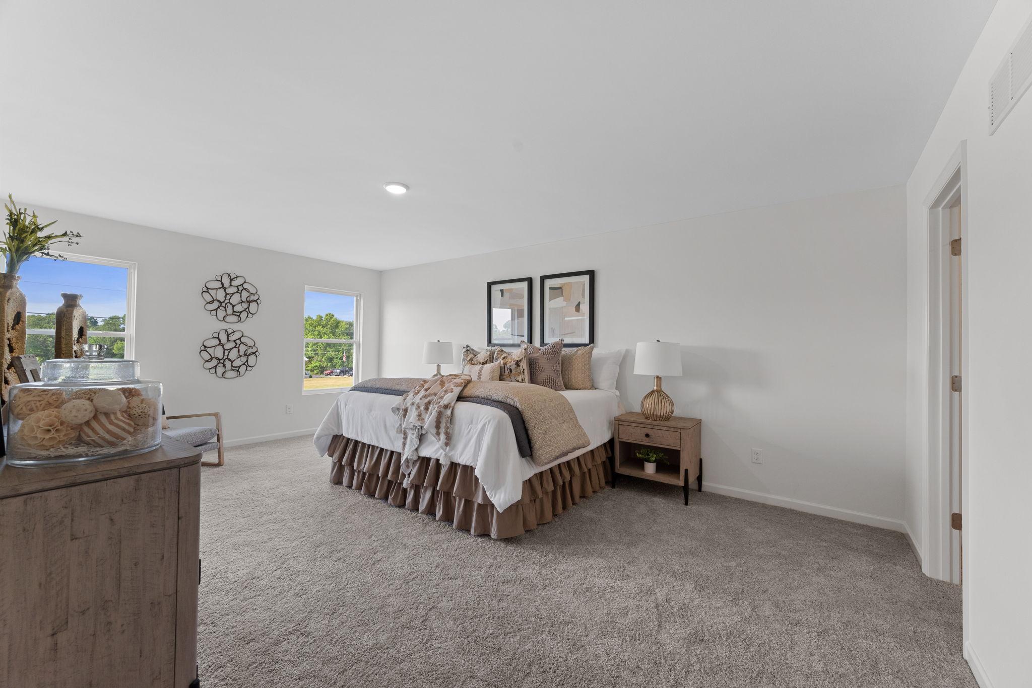 A spacious, well-lit bedroom featuring a neatly made bed with decorative pillows, bedside tables with lamps, large windows providing natural light, and wall art accents adding a cozy and modern touch.