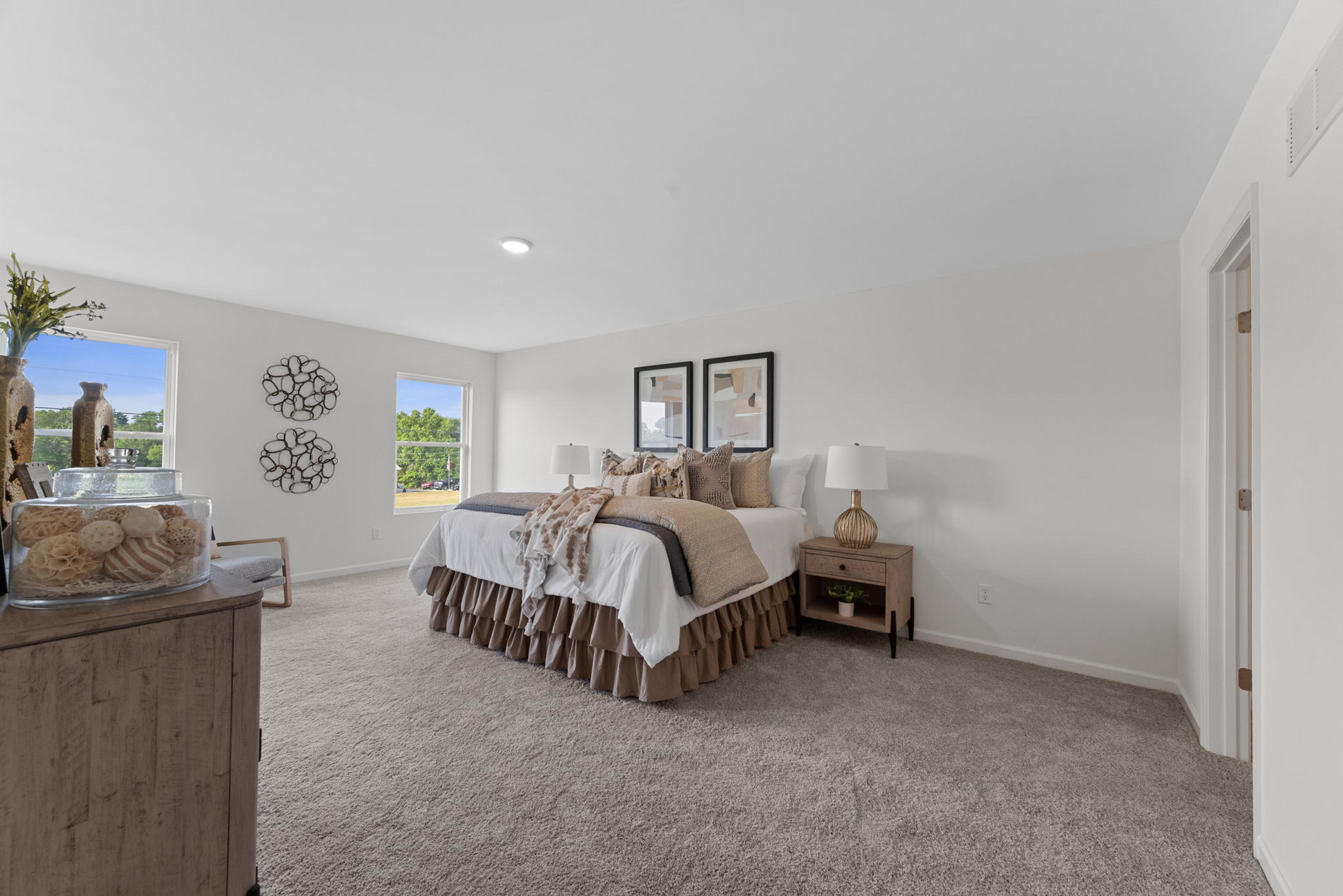 A spacious, well-lit bedroom featuring a neatly made bed with decorative pillows, bedside tables with lamps, large windows providing natural light, and wall art accents adding a cozy and modern touch.