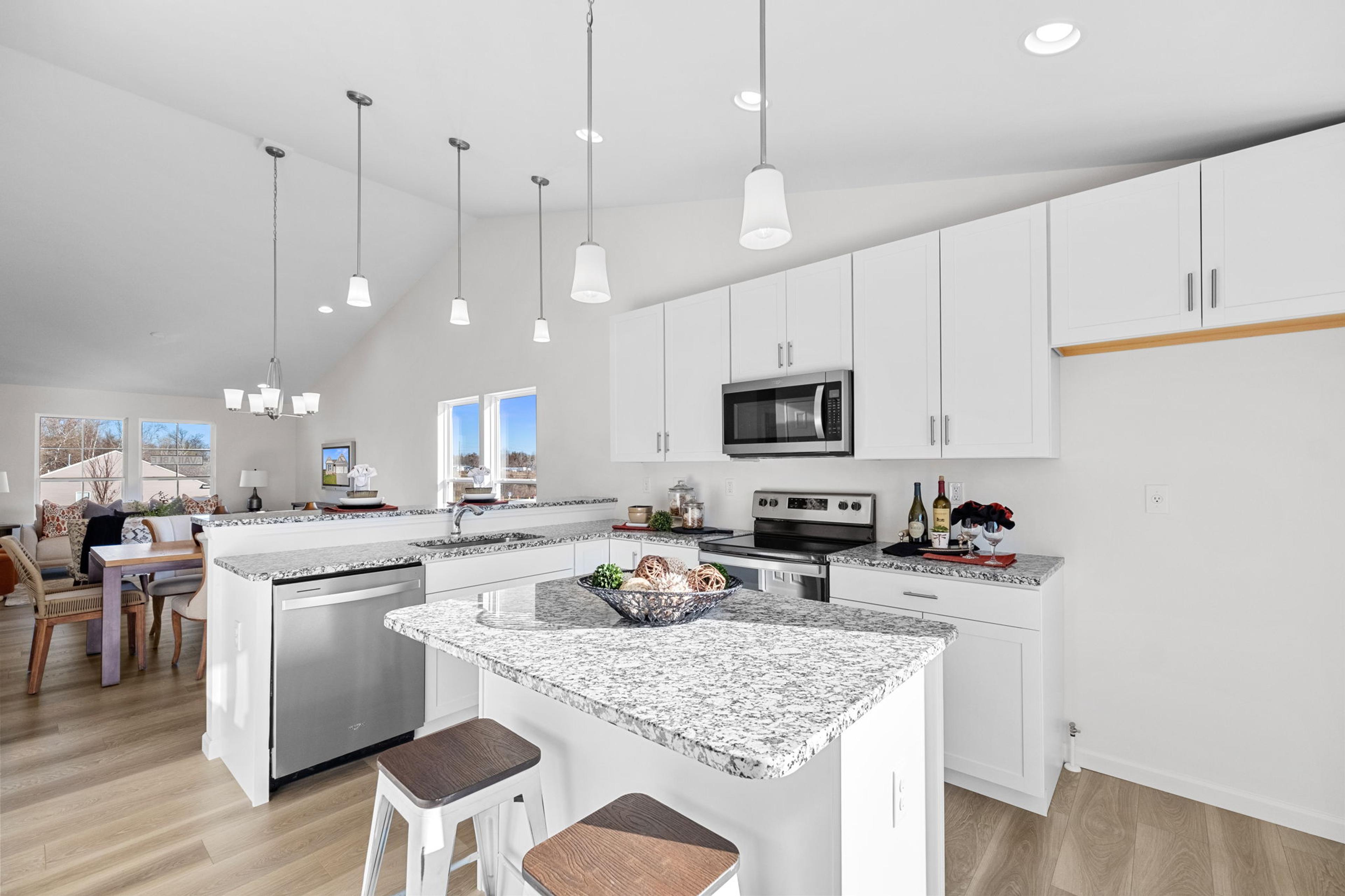 A contemporary kitchen with white cabinets, granite countertops, and stainless steel appliances, centered around a spacious island with seating. Bright pendant lights and an open layout make the space modern, functional, and inviting.