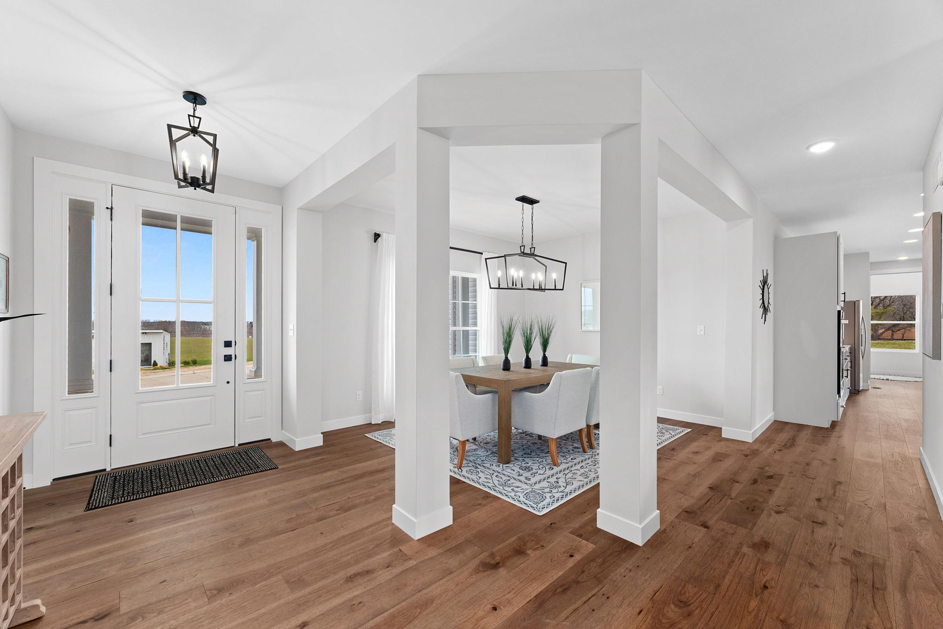 A bright and airy entryway with double doors, elegant light fixtures, and rich hardwood flooring, leading to an open dining area framed by white columns. The space combines modern design with a welcoming, functional layout.