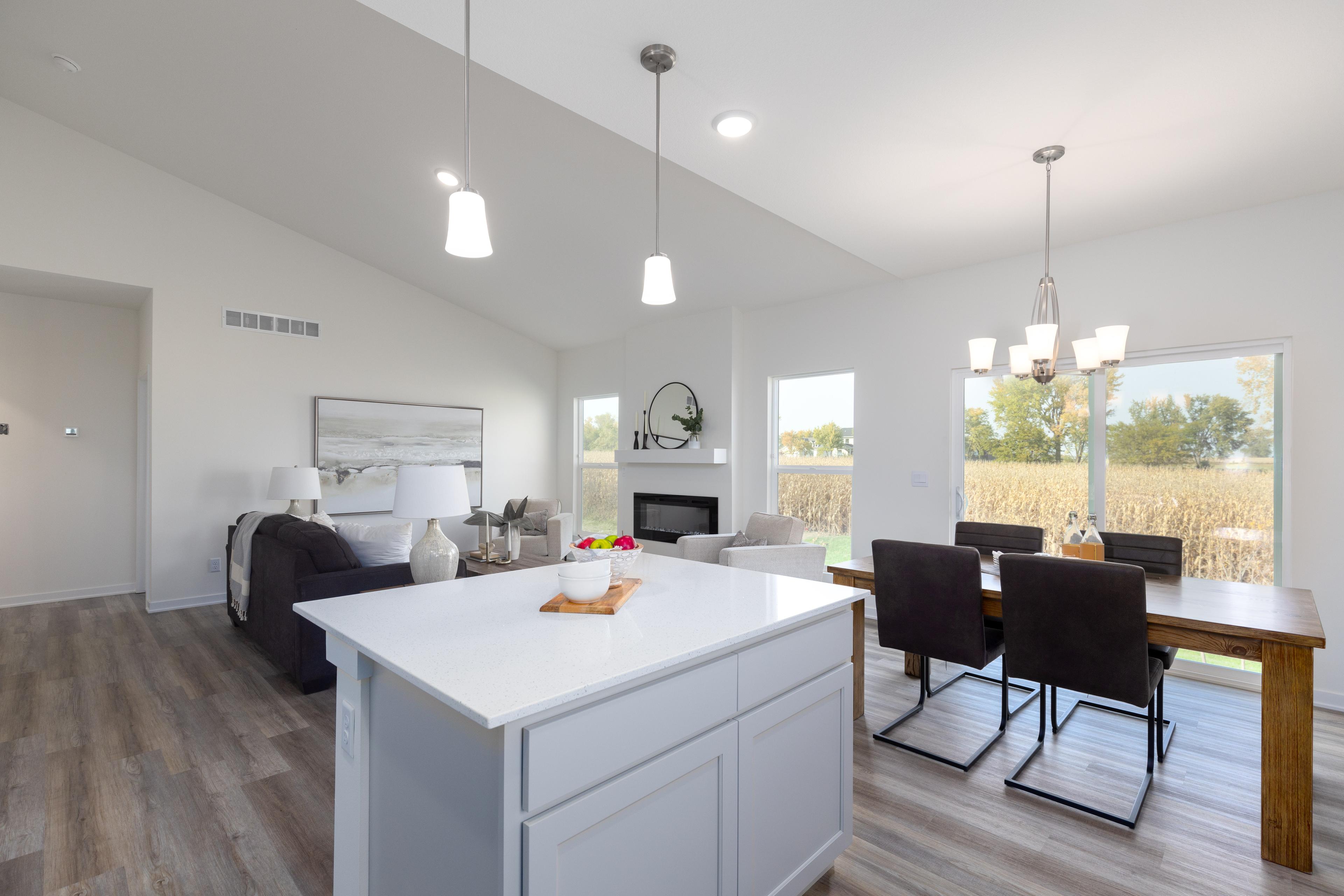 A bright and open-concept living space with a modern kitchen island at the forefront, featuring sleek white cabinetry. The area seamlessly transitions into a cozy living room with a fireplace and large windows that allow natural light to fill the room, highlighting the dining area with a view of the outdoors.