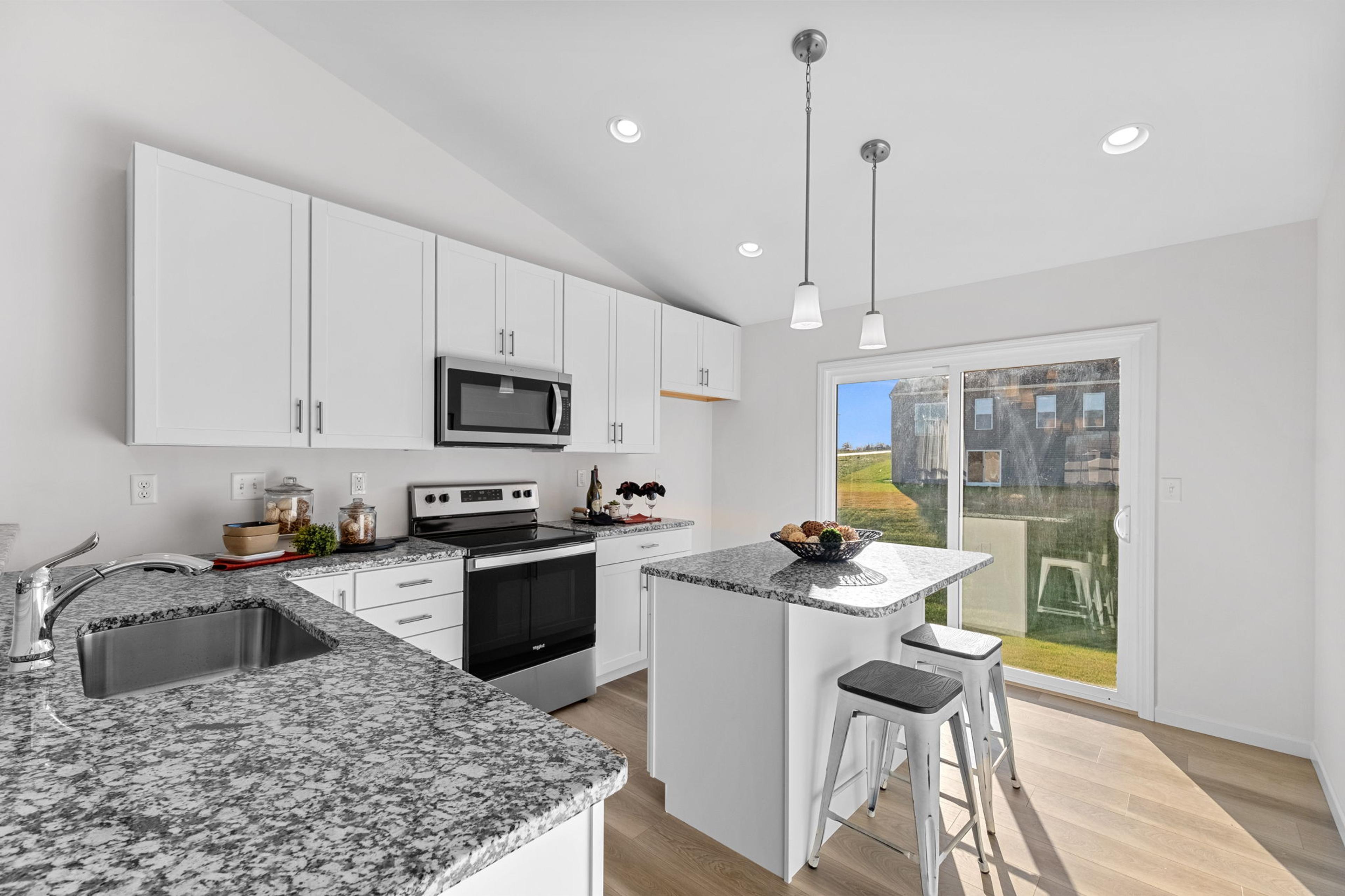 A bright, modern kitchen with white cabinets, granite countertops, stainless steel appliances, and an island with pendant lighting and bar stools, opening to a patio through sliding glass doors.