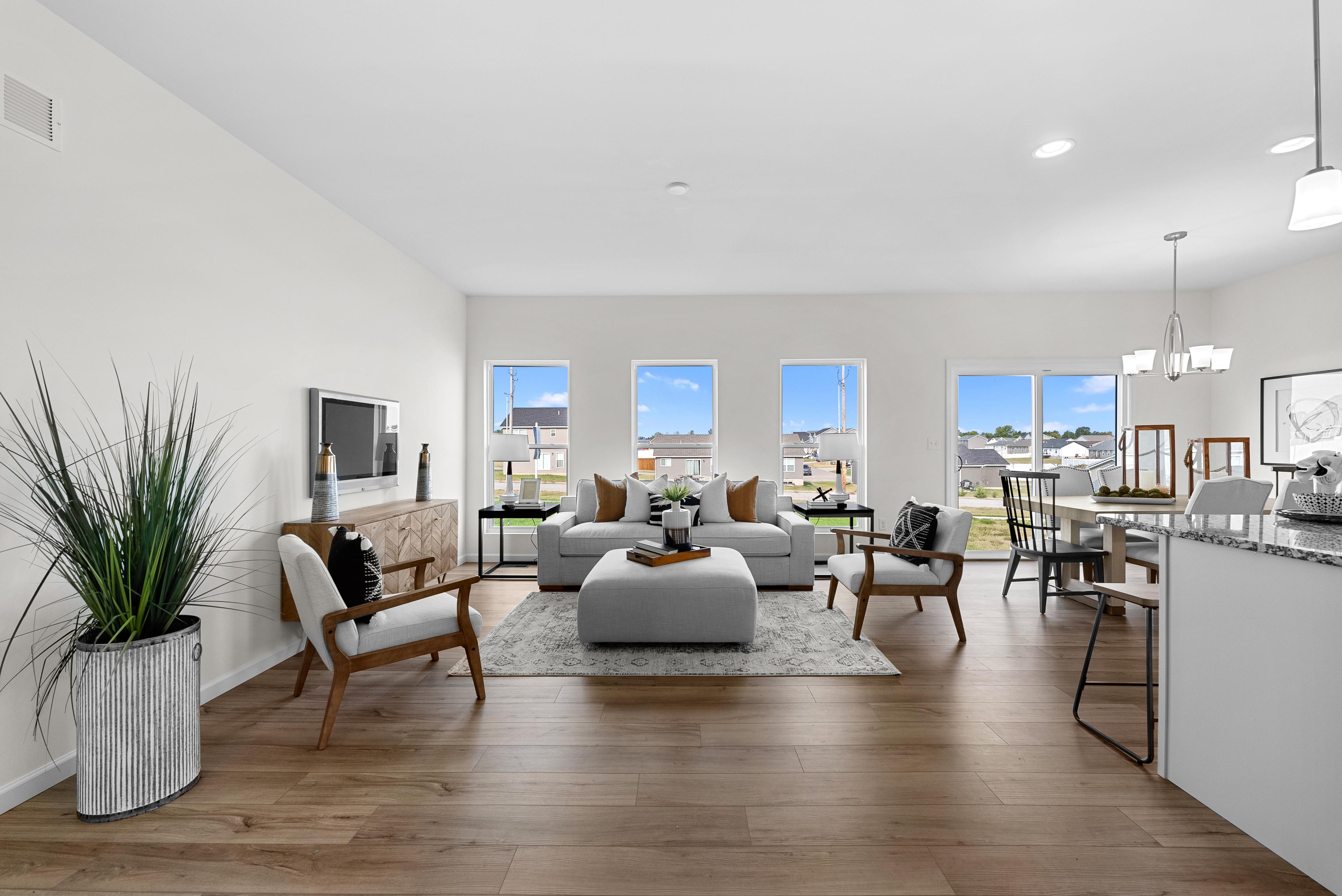 “Bright and airy living room featuring large windows, modern furniture, and wood flooring, seamlessly flowing into a dining area and kitchen for an open-concept design.