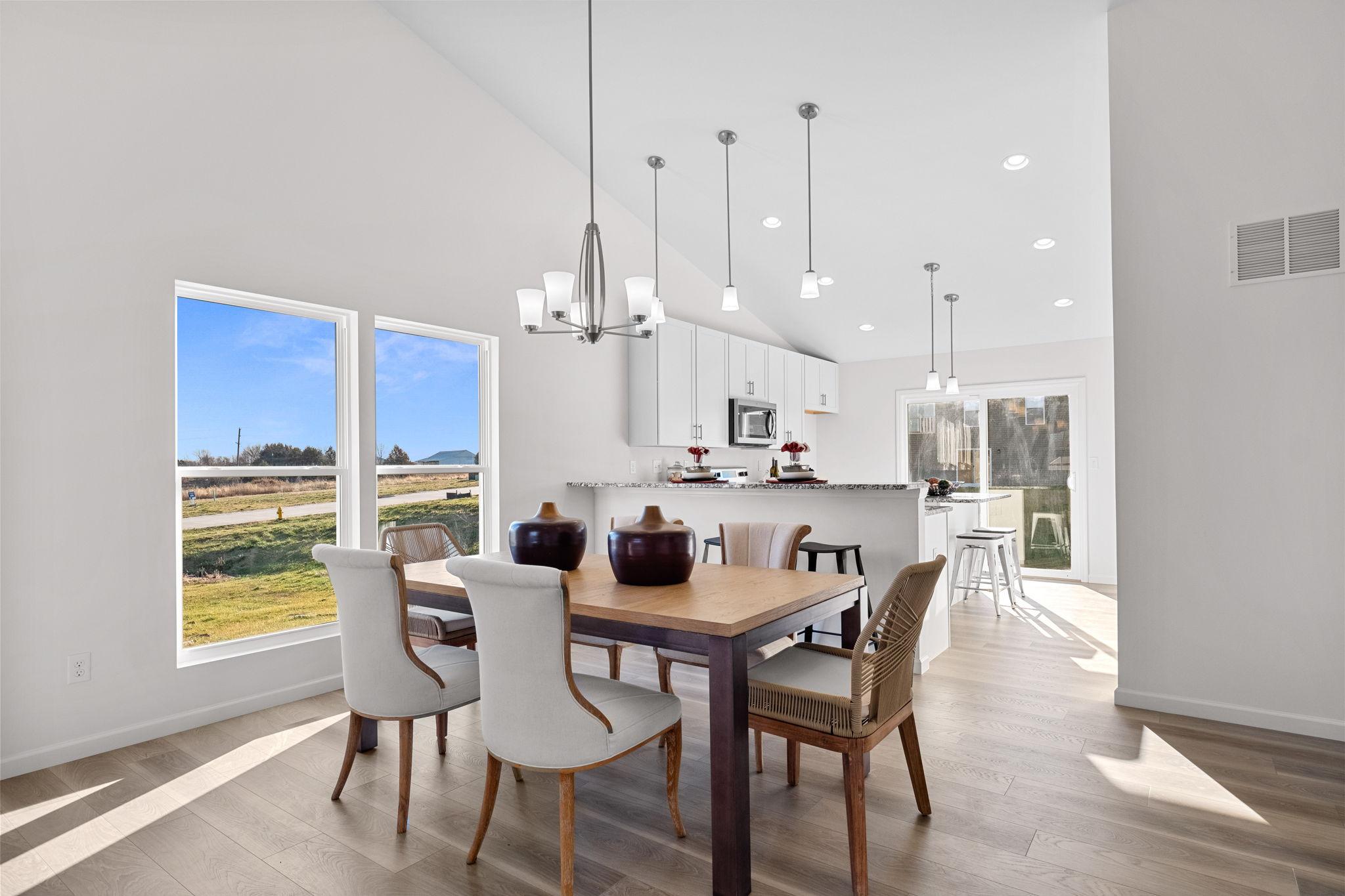 A bright, modern dining area with a wooden table, elegant chairs, and a chandelier, adjacent to a sleek white kitchen featuring pendant lights, large windows, and a view of the outdoors.