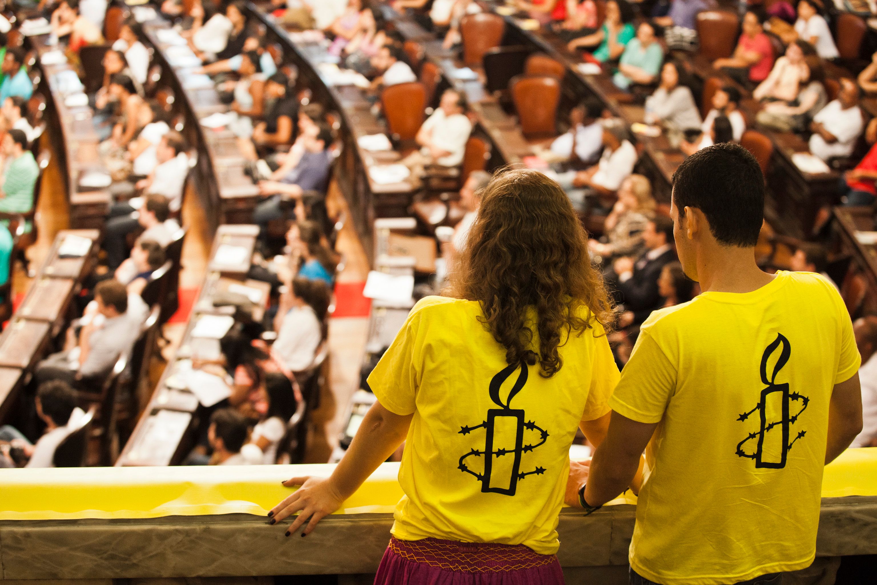 Two people with Amnesty International logos on their t-shirts looking down at a room of people.