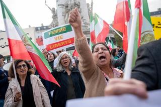 Three women stand at a protest, holding flags and signs, and they are shouting.