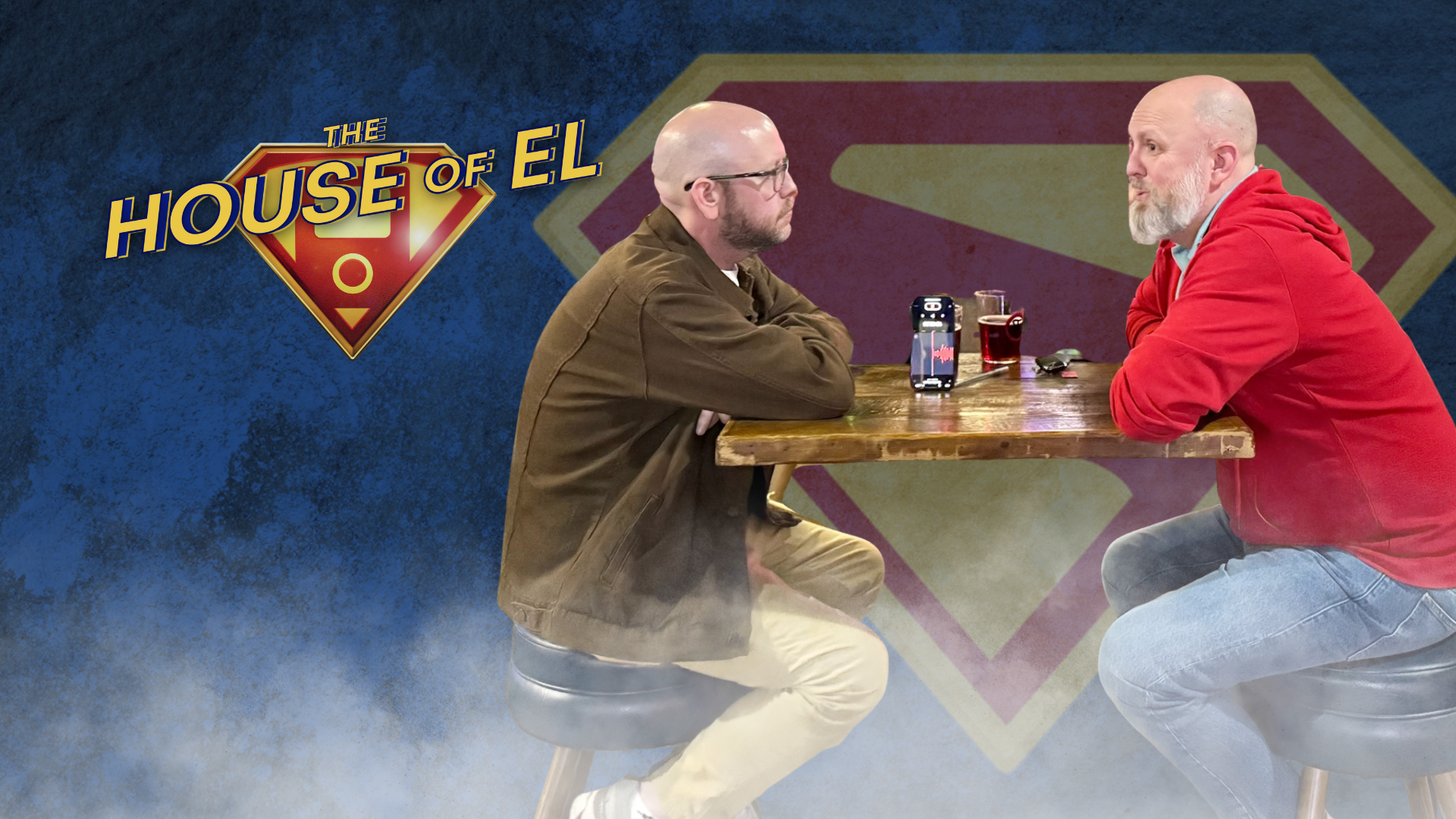 Two men sit at a table in front of a Superman logo.
